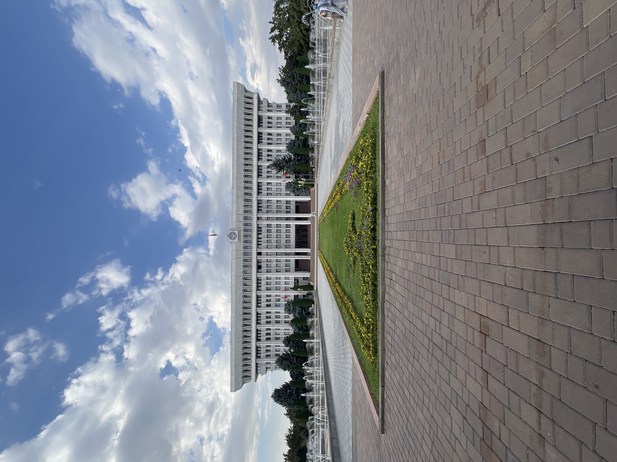 The image captures the Ala-Too Square in Bishkek, Kyrgyzstan, featuring the White House (Presidential Palace) as the central subject. The imposing, multi-story white building with numerous windows and ornate columns dominates the background. In front of the building, a large plaza is laid with interlocking brick pavers, leading towards a manicured green lawn bordered by flowerbeds with yellow and red blooms. Beyond the lawn, a long, rectangular fountain system with multiple jets sprays water upwards. Tall, dark green coniferous trees line the sides of the square, and a blue sky with scattered white clouds forms the backdrop. In the lower right portion of the frame, three people, two women and a man, are walking away from the camera. The scene is devoid of any overt political activity or protests, presenting a calm and public space.