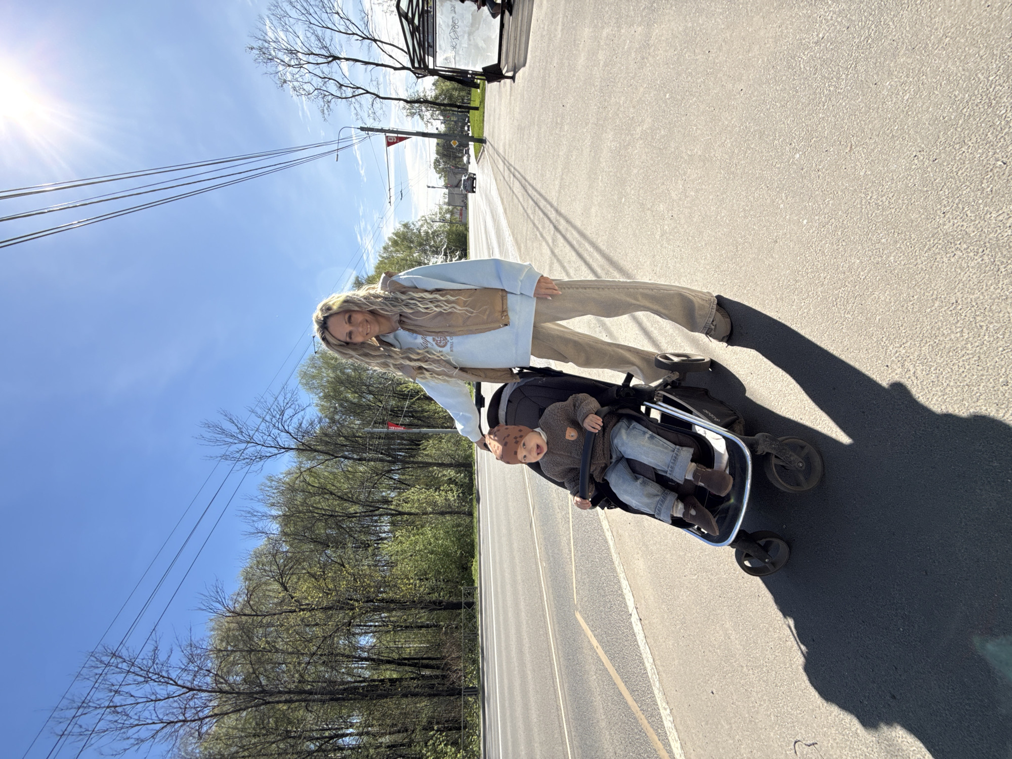 A woman is pushing a baby in a stroller on a paved road in an urban environment. The woman has long blonde hair, is wearing a light blue hooded sweatshirt, a brown vest, and tan pants. The baby in the stroller is wearing a brown outfit and a hat. They are on a road with painted lane markings and trees lining the side of the road. Overhead electrical wires are visible against a blue sky with the sun shining brightly. A bus stop shelter is also visible in the background.