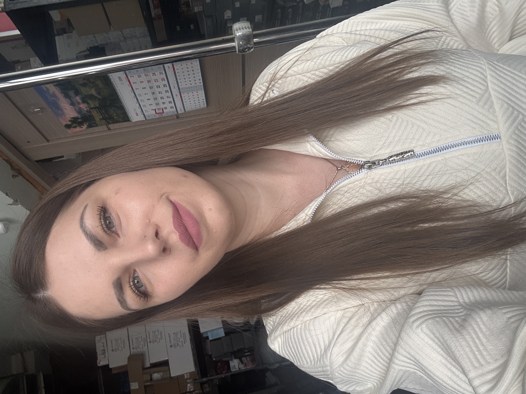A woman with long brown hair is pictured wearing a textured white zip-up jacket. She has fair skin, well-defined eyebrows, and her lips are painted with a muted pink lipstick. In the background, a calendar is visible, possibly attached to a shelf or cabinet. Beneath the shelf, there are multiple cardboard boxes. The scene does not explicitly confirm the location is in Klin, Russia.