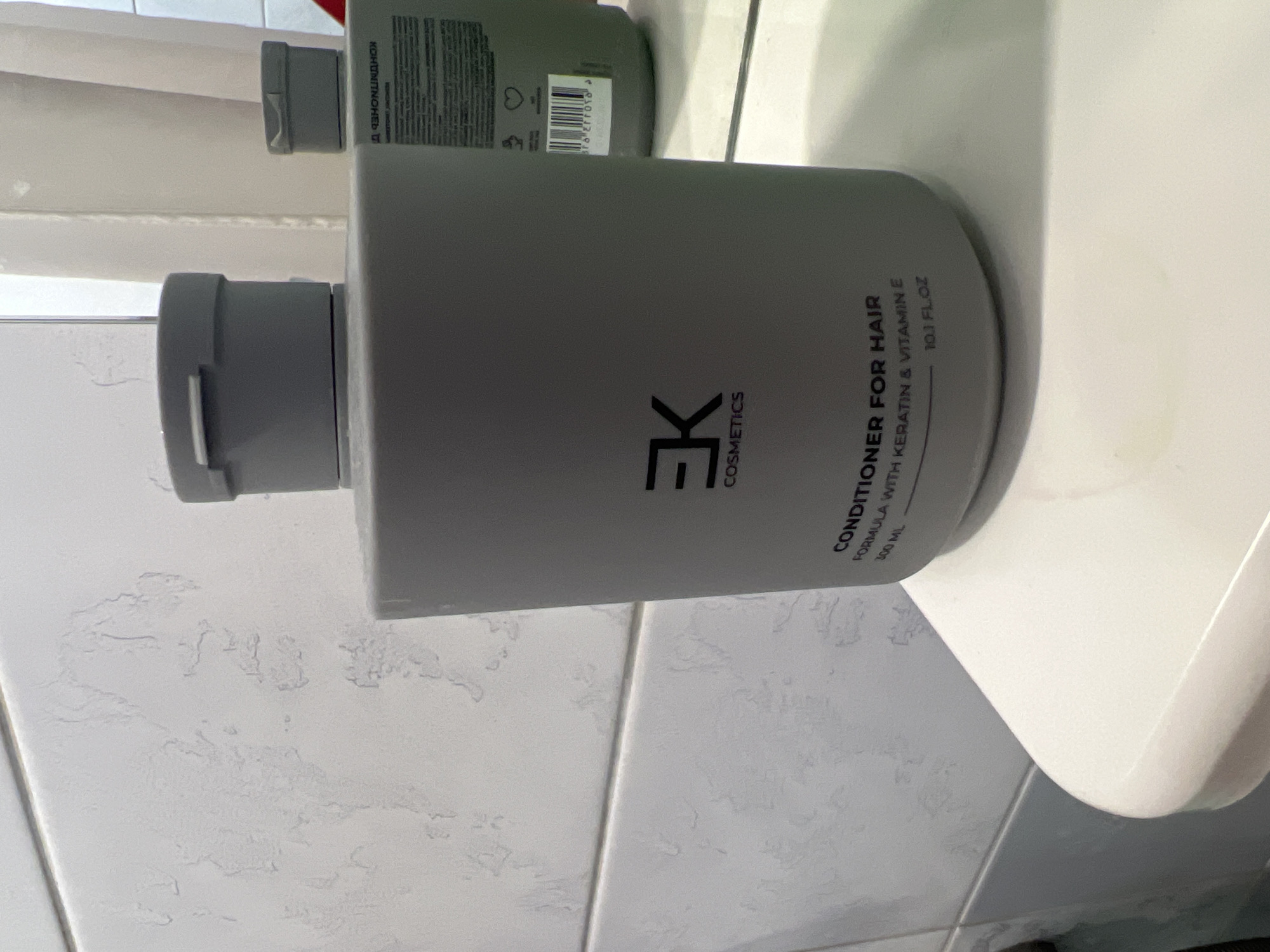 The image shows a grey bottle of "CONDITIONER FOR HAIR FORMULA WITH KERATIN & VITAMIN E 300 ML" on a white surface. The bottle has a black logo that appears to be "EK COSMETICS". In the background, there is a tiled wall and another bottle with text in Russian. The scene appears to be a bathroom. There is no information in the image to determine its location in the city of Ulyanovsk, Russia.