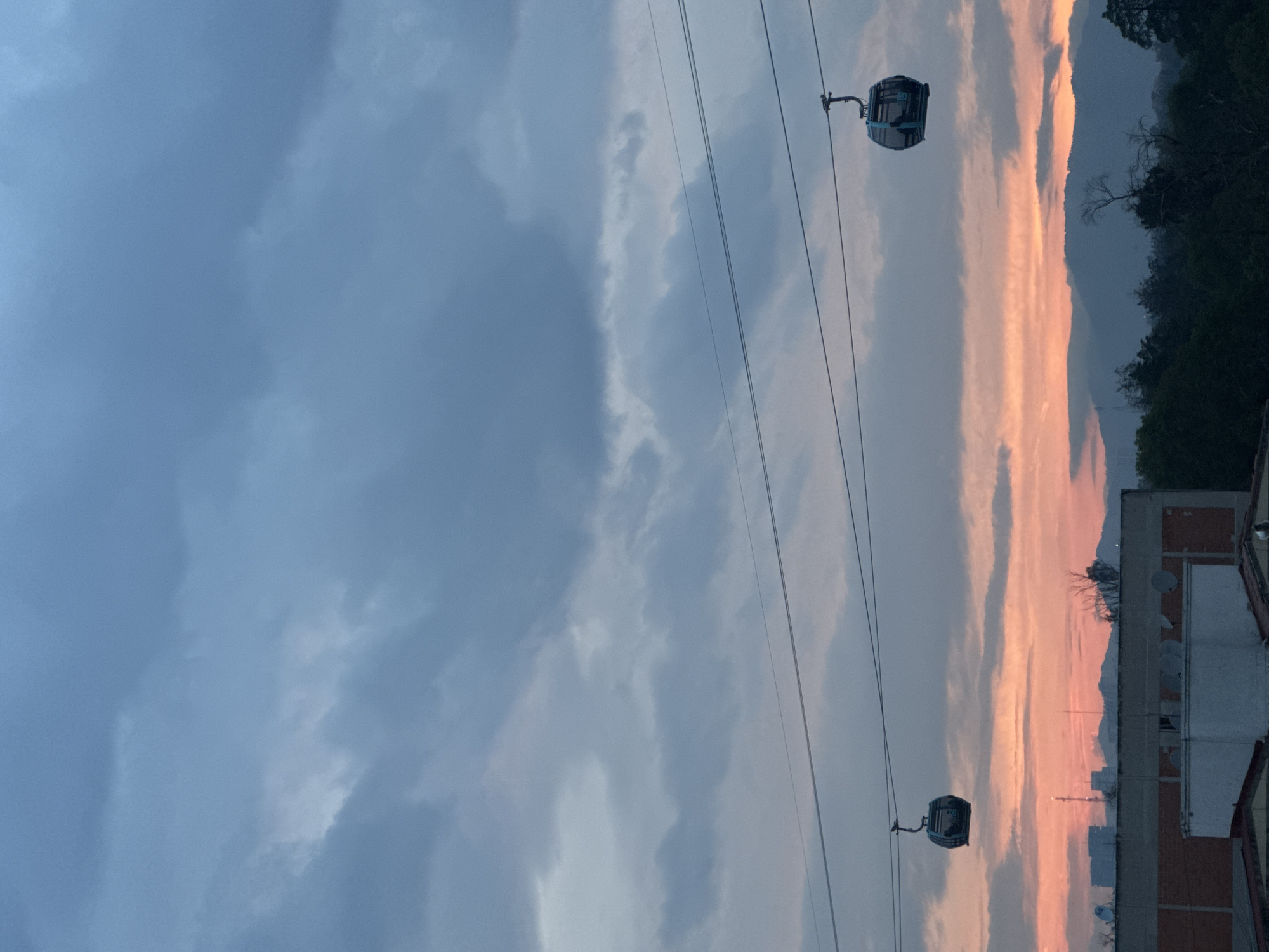 The image depicts a cityscape at dusk, featuring a cable car system. Two cable cars are visible, suspended from overhead cables. The sky is characterized by a mix of blues and pinks, suggesting a sunset or sunrise. A building is partially visible at the bottom of the frame, along with trees on the horizon. The location is Unknown, Unknown.