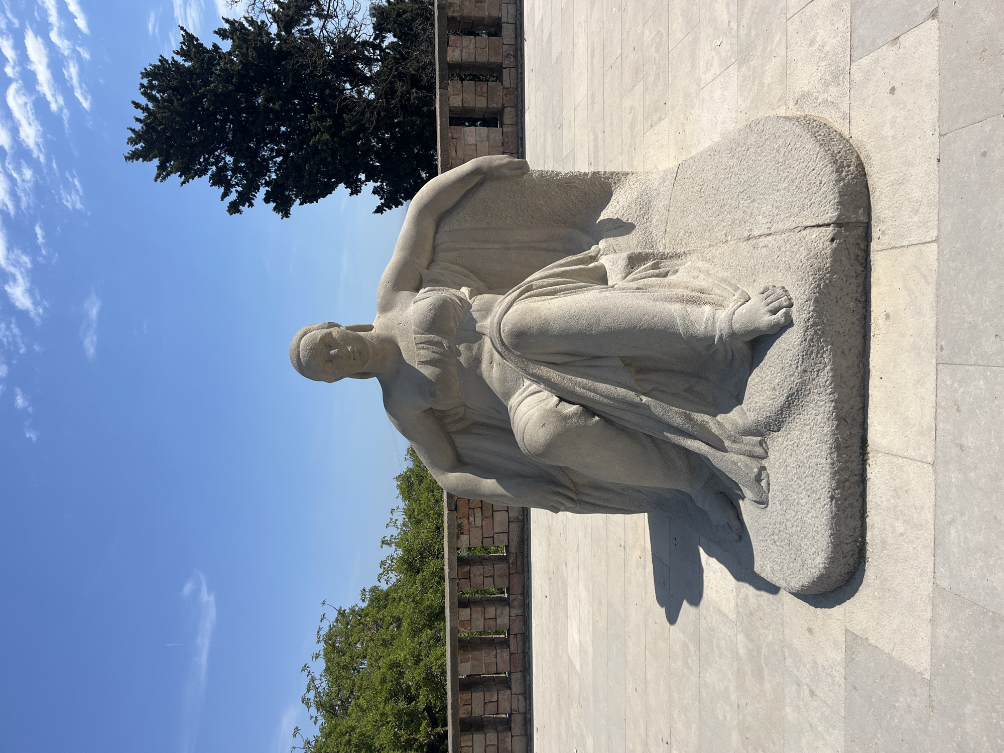 The image shows a stone sculpture of a reclining woman. The sculpture is positioned on a stone platform. In the background, there is a section of a stone wall with greenery visible behind it, a tree, and a blue sky with clouds. The sculpture is located in Barcelona, Spain, most likely in a park or historical site.