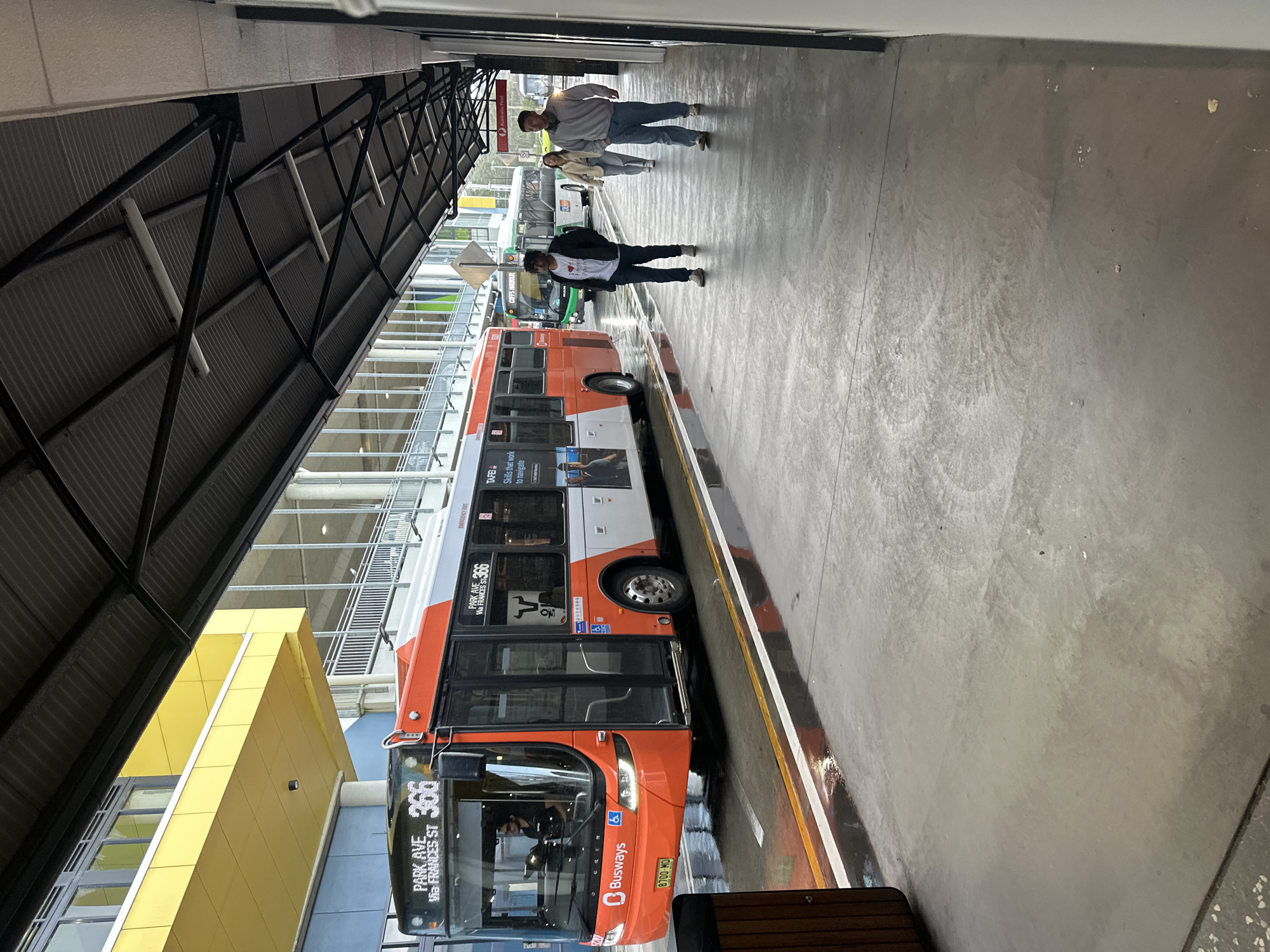 This is a medium-angle, outdoor, eye-level shot of a bus station in Coffs Harbour, Australia, under a covered walkway. The foreground is dominated by a wet concrete pedestrian walkway, with a series of light grey circular patterns visible on the surface. To the left, a dark wooden bin is partially visible.
In the mid-ground, an orange and white Busways bus, route number 366, is stopped at a bus bay. The bus displays "PARK AVE Via FRANCES ST" on its digital destination board. A driver is visible in the driver's seat. Next to the bus, on the walkway, three individuals are walking away from the viewer. The closest is a young man wearing a black hoodie and dark jeans. Behind him, a young woman in a light-colored jacket and light blue jeans walks, followed by a man in a grey hoodie and light blue jeans. Further in the background, another green and white bus is parked.
The background features a modern building with yellow and grey architectural elements. The covered walkway above the bus bay has a dark, corrugated metal roof supported by dark metal beams. The overall lighting suggests an overcast day.