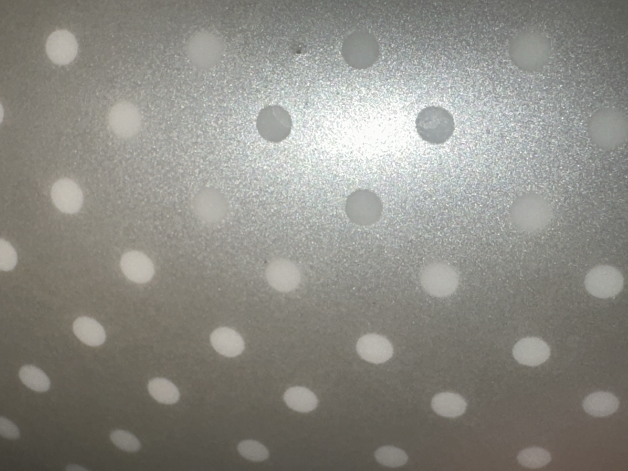 The media file is a close-up, abstract photograph of a textured, metallic surface with a pattern of evenly spaced white dots. The surface appears to be a light gray or silver color, with a slightly rough or matte finish that catches the light in places, creating a subtle shimmer. The white dots are circular and appear to be slightly raised or printed onto the surface, with soft edges that suggest they might be slightly blurred or illuminated. The perspective is angled, giving the impression that the surface is curved or viewed from an oblique angle. There is a bright light source in the upper right quadrant of the image, which causes some of the dots in that area to appear more luminous and even slightly washed out. No people, objects, or discernible scene are visible, only the repetitive geometric pattern. The image is abstract and lacks specific details about time of day, weather, or location, other than the provided context of Letterkenny, Ireland. There is no visible text within the image.