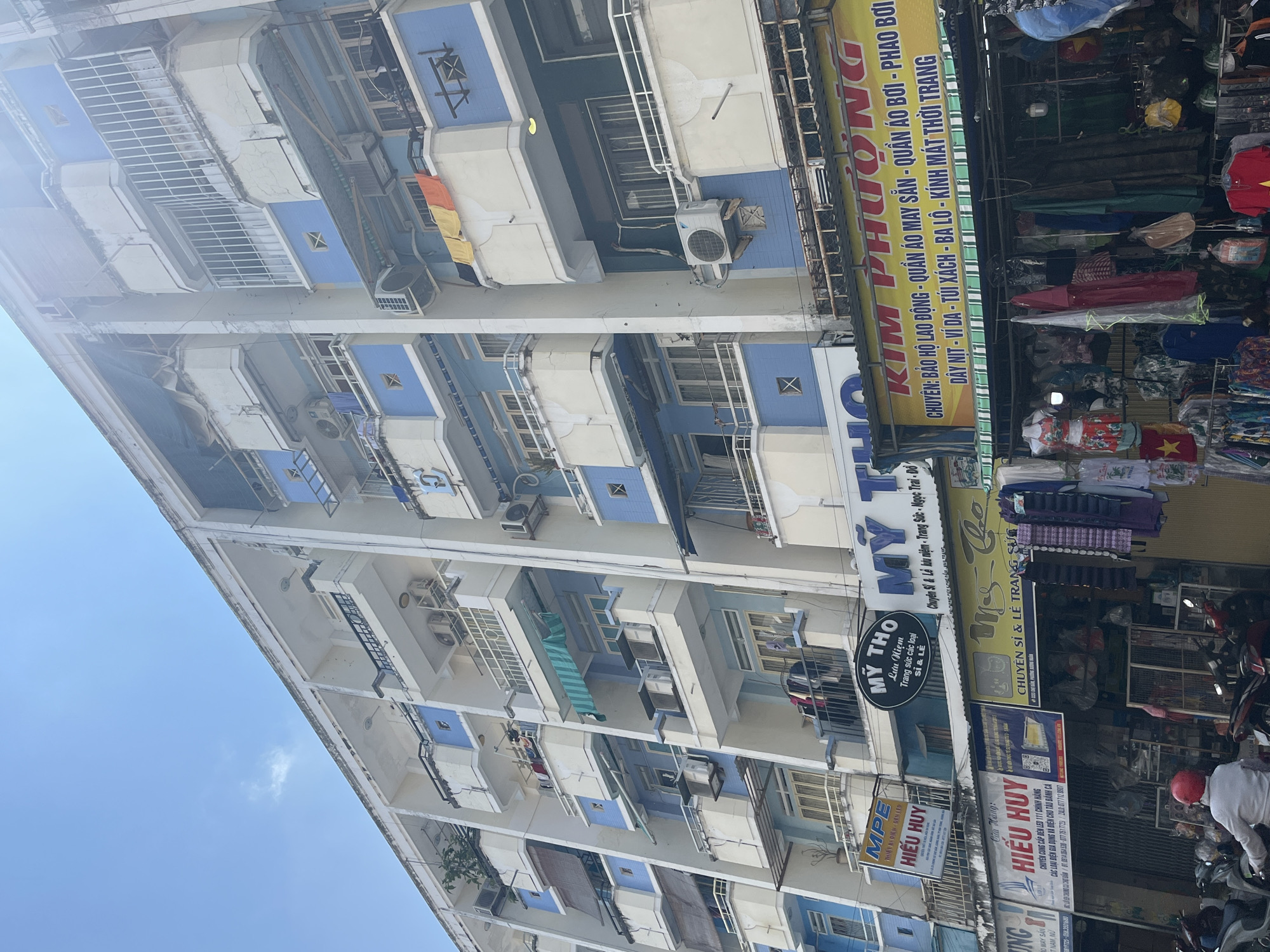 The image shows a multi-story building in Nha Trang Ward, Vietnam. The building is light blue and white with balconies. Several shops are visible at the ground level, with signs displaying business names like "HIẾU HUY," "MỸ THO," and "KIM PHƯỢNG," offering products from LED lights and souvenirs to clothing. There is a motorbike parked in front of one of the stores, a person on the motorbike.