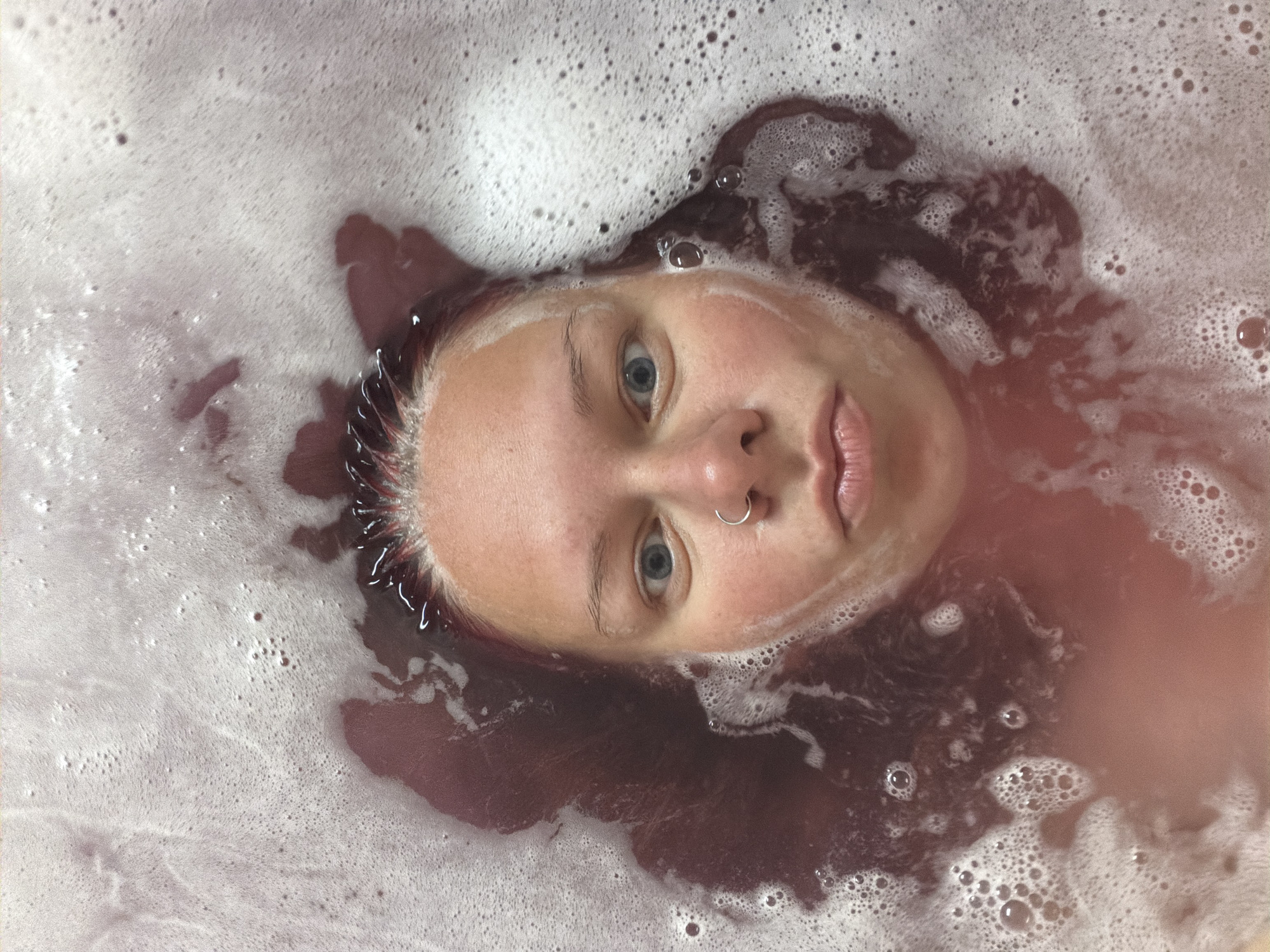 A close-up, top-down shot shows a person's face emerging from a bath filled with reddish-purple water and white bubbles. The subject has light skin, blue eyes, and dark hair. They are wearing a silver hoop nose ring. The lighting is soft, and the focus is on the face, with the surrounding water and bubbles creating a soft, diffused effect. The background is indistinct due to the water and bubbles. There is no identifiable information in the image that would suggest a specific location within the city of Chaikovsky, Russia.