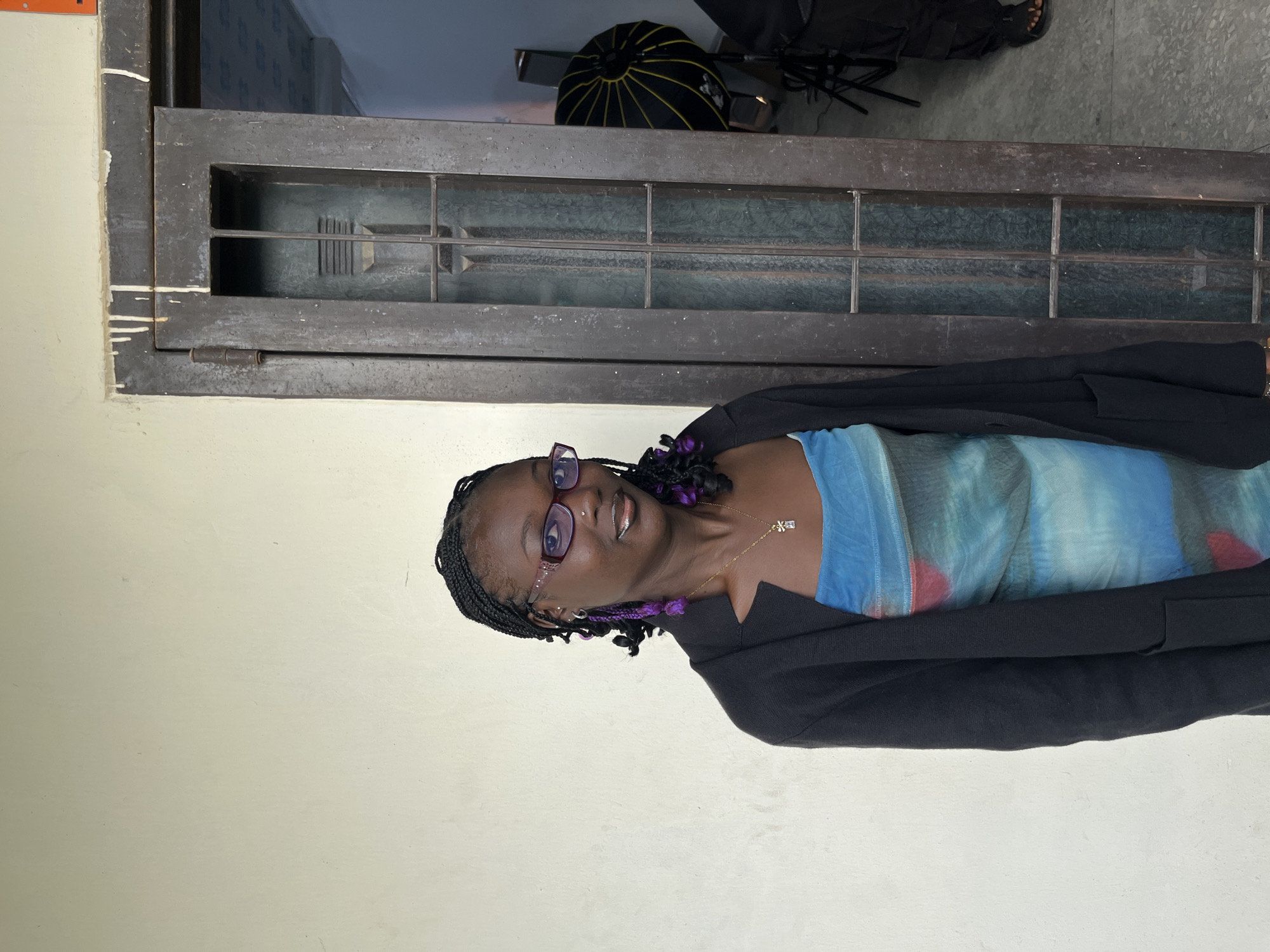 A Black woman with braided hair and purple accents stands against a textured wall, facing the camera. She wears purple-rimmed glasses, a black cardigan over a blue and green tie-dye dress, and a silver necklace with a pendant. She is positioned to the left of a dark wooden door with a glass pane divided by metal bars. To the right of the door, a black umbrella-shaped photography light is visible. The environment suggests an indoor or semi-covered space, possibly a studio or entryway, with a concrete floor. The overall scene is well-lit, with soft lighting.