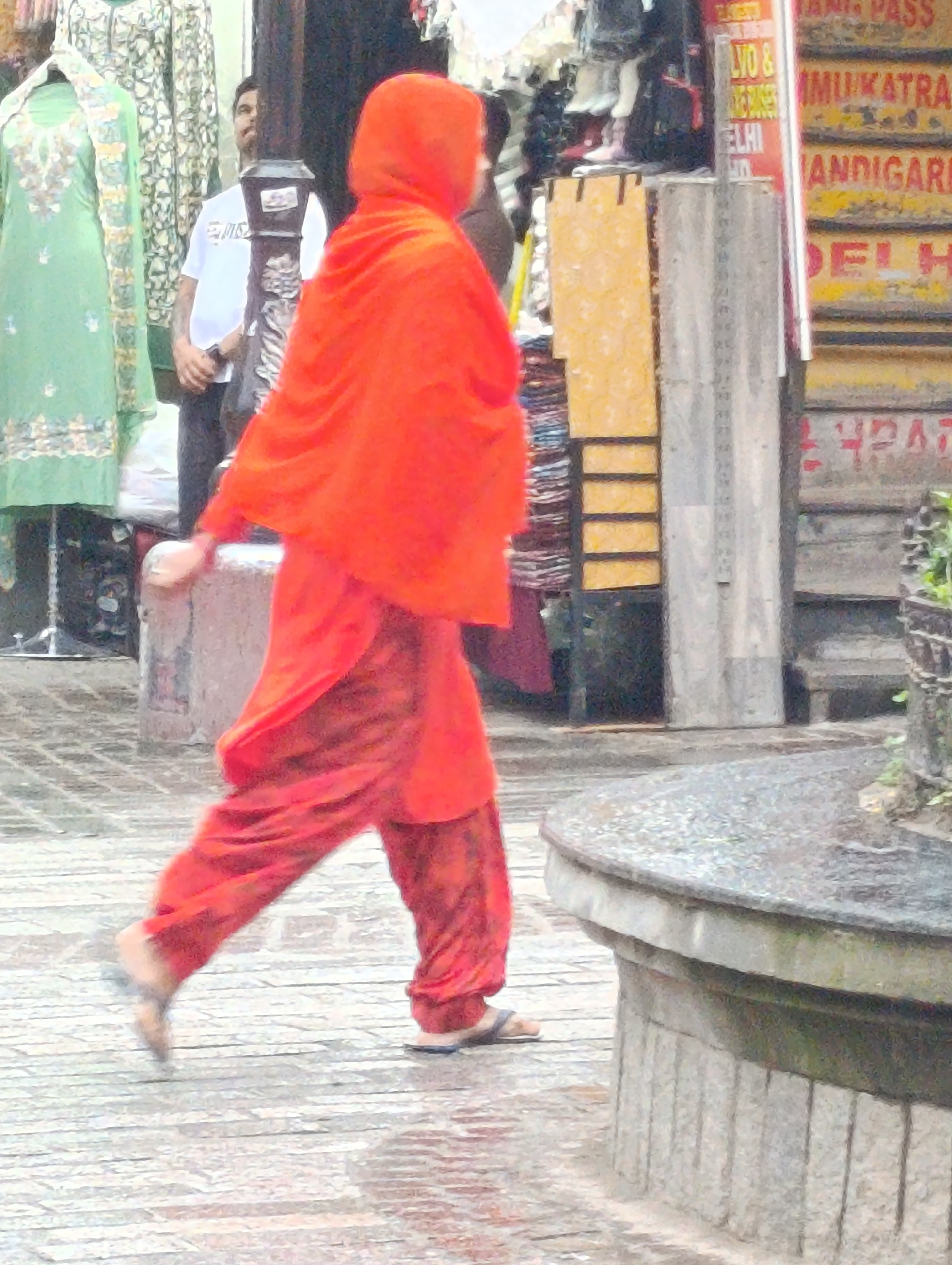 A person in an orange outfit walks on a paved street, partially obscuring a stall with yellow and grey shelves. The person's left foot is barefoot, and their right foot wears a sandal. To the left, a green dress is displayed on a mannequin, and a man in a white t-shirt stands behind it. In the background, signs with red lettering are visible on a building. The scene appears to be a street market in Manali, India.