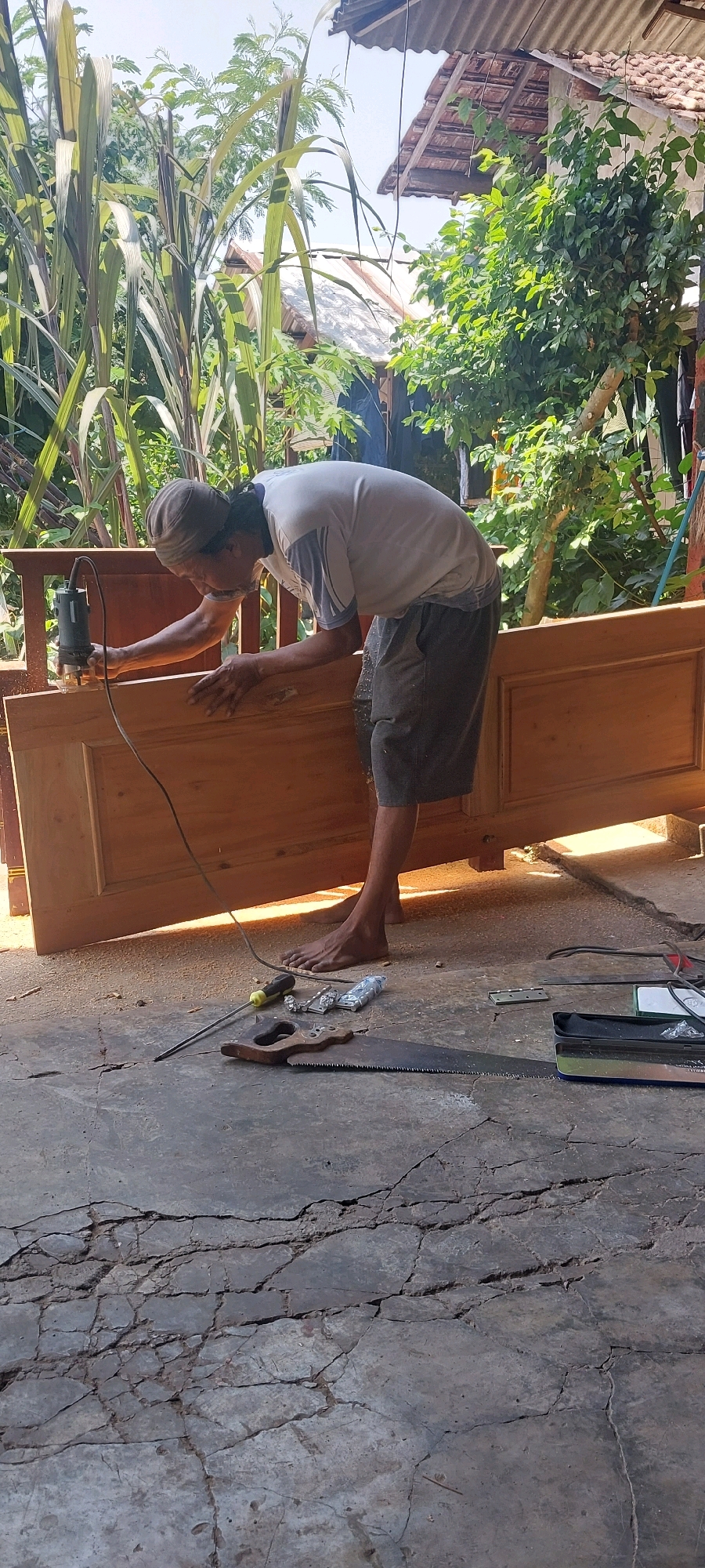 In Keling, Indonesia, an elderly man with a headscarf is using a router to shape a wooden door. He is barefoot and wearing a grey t-shirt and shorts. He is standing in what appears to be a workshop or outdoor area, with a cracked concrete floor covered in sawdust. Various woodworking tools are scattered around, including a saw, screwdriver, and hinges. In the background, lush green foliage and a building with a tiled roof are visible, suggesting a rural or semi-rural setting. The sunlight casts strong shadows, indicating it is daytime.
