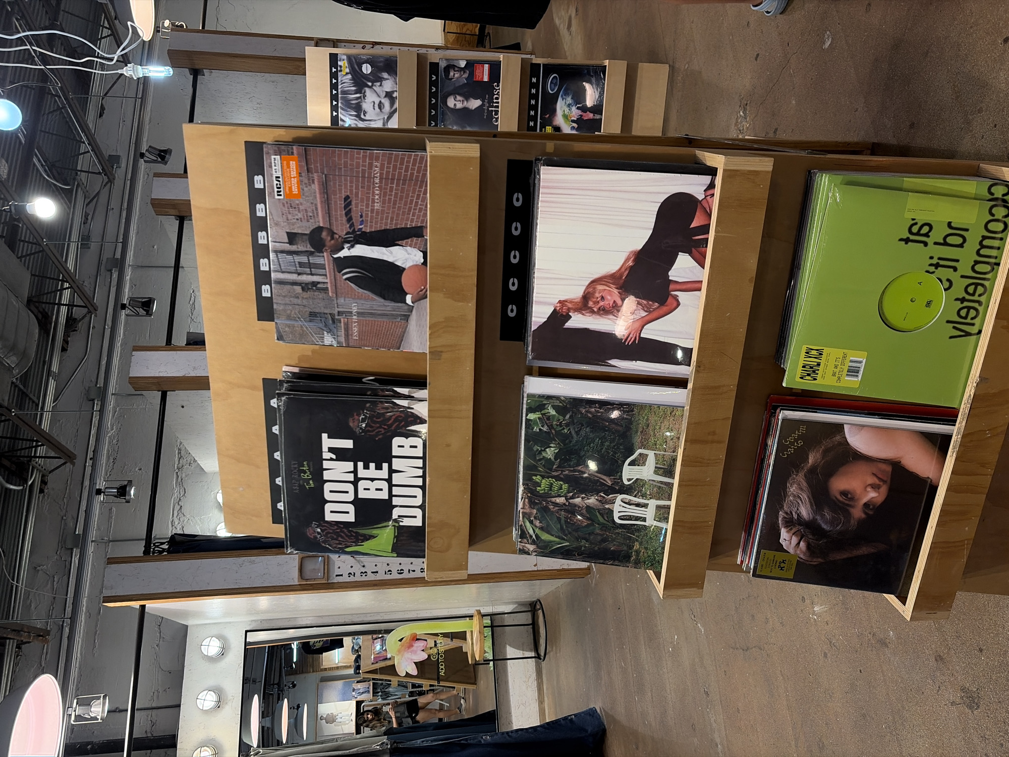 The image shows several shelves filled with vinyl records, likely in a store. The records feature various artists and album covers, including A$AP Rocky's "Don't Be Dumb", Blood Orange's "Essex Honey", and Charli XCX's "Brat And It's Completely Different". Other albums include "twilight eclipse." The shelves are made of light wood and are arranged in a tiered fashion.
The setting appears to be a retail environment, given the arrangement of the records and the presence of store fixtures in the background, including a clothing rack and a mirror with illuminated light bulbs. Given the reflections of clothing and other retail items, it's probably a clothing store in addition to selling records. Based on the reflections and store fixtures, it may be in the city of Austin, United States of America.