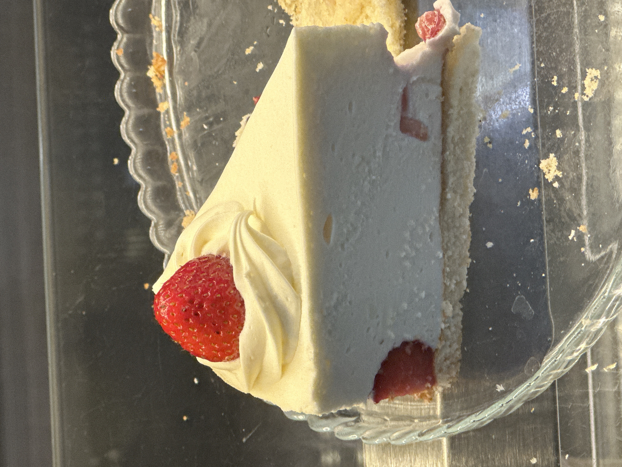 A close-up, slightly overhead shot presents a triangular slice of a creamy white dessert, likely cheesecake or mousse cake, resting on a clear, scalloped glass plate. The dessert slice features a distinct crust or base layer, light brown in color, forming the bottom and back edge of the slice. The main body of the dessert is a smooth, off-white or pale cream color, with small red fruit pieces (appearing to be strawberries) visible within it, particularly towards the thinner end and embedded near the top edge.
At the wider end of the slice, a swirl of pale yellow whipped cream is piped, partially cradling a fresh, vibrant red strawberry. The strawberry is whole and firm, with visible seeds and a slightly textured surface.
The glass plate beneath the dessert is partially covered in crumbs and small fragments of the crust, suggesting some prior movement or cutting. The background is dark and somewhat reflective, creating a contrast with the bright dessert and indicating the possible setting of a display case or counter. No people or text are visible in the image. The lighting appears to be from above or the front, highlighting the texture of the whipped cream and the strawberry. The overall impression is one of a single portion of a dessert ready for consumption.