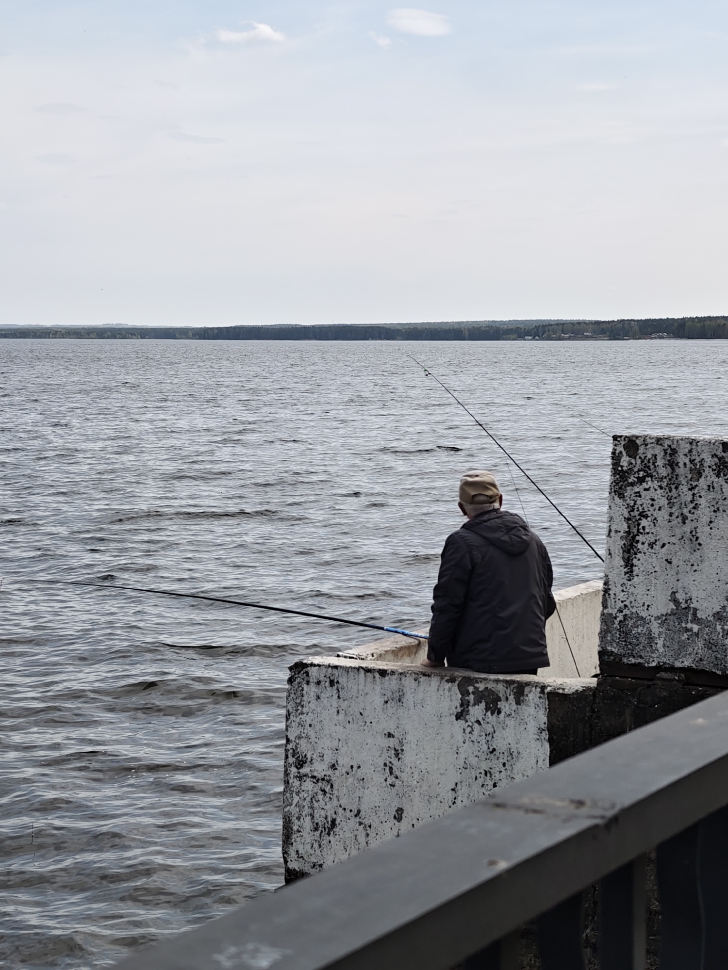 An eye-level, medium shot depicts a man fishing from a concrete pier at Votkinsk Reservoir in Chaikovsky, Russia. The man, viewed from the rear, is wearing a dark jacket and a light-colored baseball cap. Two fishing rods extend into the water, one angled low and the other reaching diagonally upward. The water surface is choppy with gentle waves.
The pier is constructed of weathered concrete blocks, showing stains and signs of age. A dark metal railing runs across the lower right foreground of the image, partially obscuring the view. In the background, a distant treeline is visible across the reservoir under a cloudy sky.