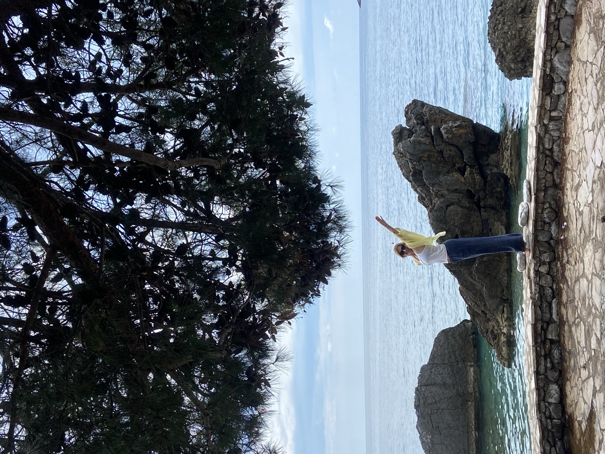 A slightly high-angle outdoor shot depicts a woman standing on a stone walkway near the sea in Prijevor, Montenegro. The image is composed of three main elements: a tree on the left, the sea with rocky outcroppings in the center, and the walkway on the right.
The woman is positioned near the edge of the stone walkway, extending one arm upwards towards the sea. She is wearing a white tank top, dark blue pants, white shoes, and a light yellow top, sunglasses.
To the left, a large tree with dense foliage dominates the view. The tree's branches and leaves create a dark, textured canopy that contrasts with the brighter background.
The sea occupies the central portion of the image, featuring a light blue color. Several rocky outcroppings are positioned within the sea, adding visual interest. The sky is partially visible in the background.
The stone walkway is constructed with light-colored stones of different sizes. A short wall made of the same stones runs along the edge of the walkway.