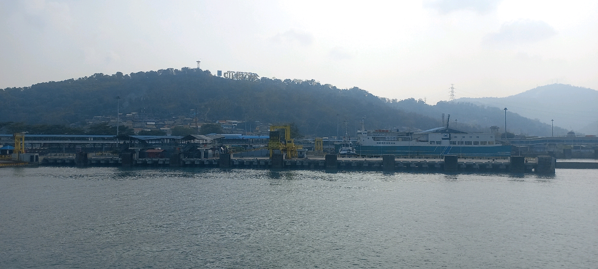The image captures a view of the Merak Port in Cilegon, Indonesia. In the foreground, the calm, grey water of the sea stretches across the frame. A concrete pier runs horizontally across the midground, with several dark, square support pillars visible. On the pier, there are various structures including a yellow crane on the left, covered walkways with visible support beams and roofs, and a large ferry docked on the right. The ferry is white with a distinct blue stripe along its side and bears the logo "JLC" on its rear. Behind the pier, a dense, green, tree-covered hillside rises, dotted with buildings and what appear to be residential structures. Prominently displayed on the hillside is a large sign with white lettering that reads "MERAK BANTEN KOTA". Power lines and transmission towers are visible on the distant hills. The sky is overcast with a pale, hazy quality. There are no identifiable individuals or specific actions taking place in the visible parts of the image.