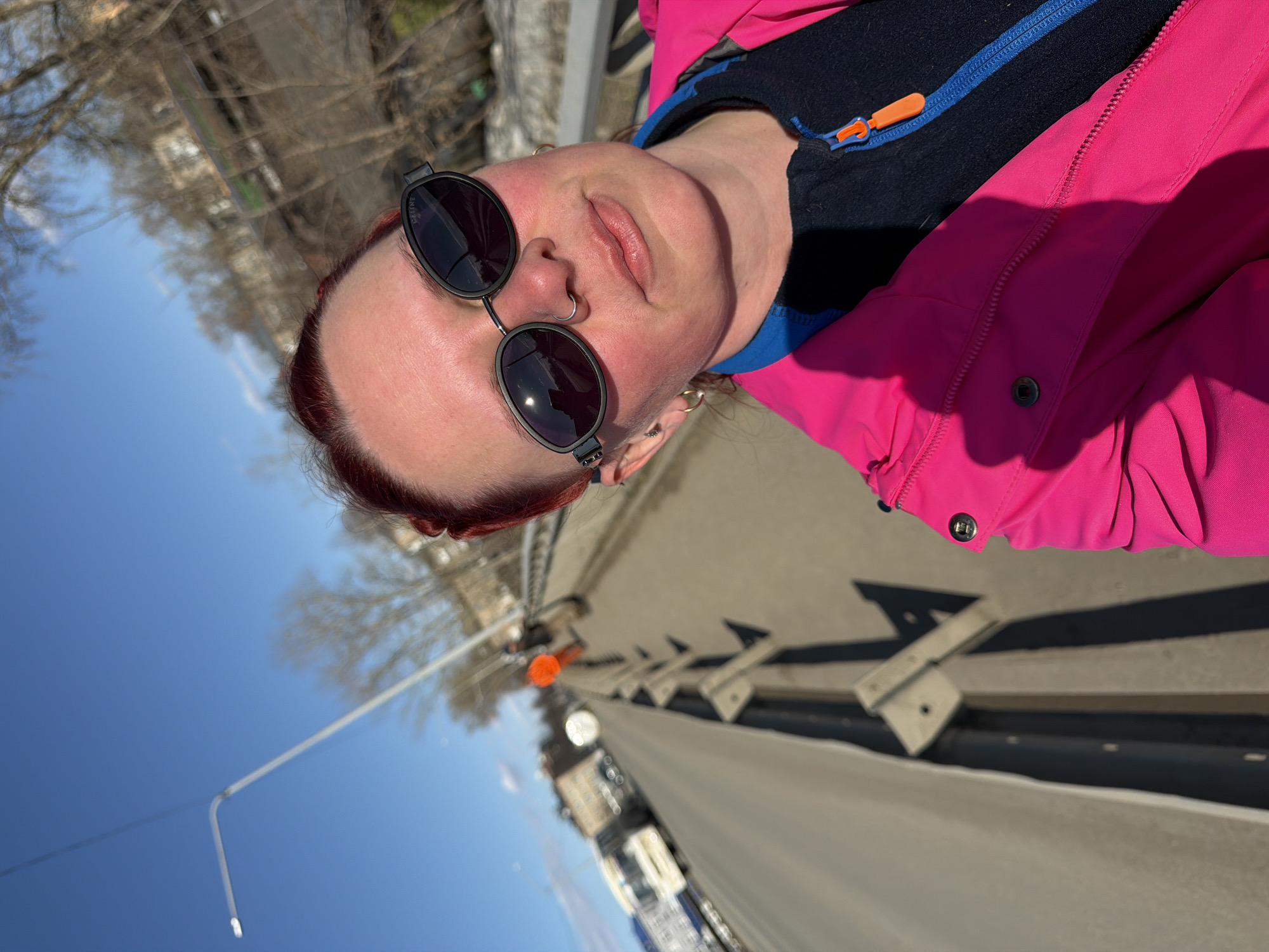 A person wearing sunglasses and a pink jacket is taking a selfie on a bridge. The bridge has concrete railings and metal supports. A street runs below. In the distance, there are buildings and trees. The sky is blue. This photo was taken in Chaikovsky, Russia.