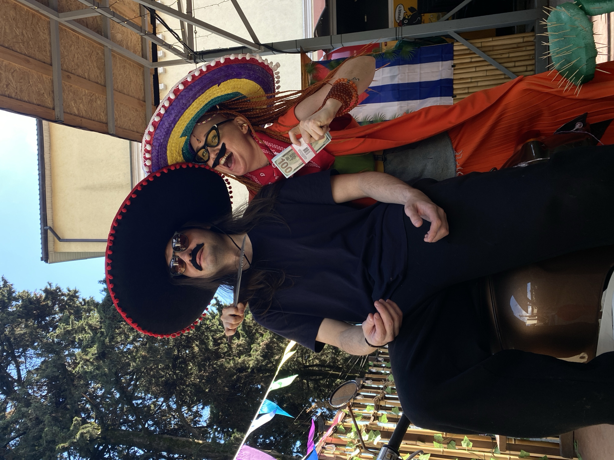 The image captures two individuals participating in a themed scene outdoors in Sochi, Russia. On the left, a male with long dark hair, wearing a black sombrero with red pom-poms, dark sunglasses, and a fake black handlebar mustache, is seated on the brown seat of a scooter or motorcycle. He is dressed in a plain black t-shirt and black pants, holding a silver-bladed knife with a dark handle pointed towards his own throat. To his right, a female with orange dreadlocks, wearing a rainbow-striped sombrero with white pom-poms, yellow cat-eye sunglasses, and a fake black handlebar mustache, stands leaning in. She is dressed in an orange top over a grey vest, a red bandana around her neck, and beaded bracelets on her right wrist. Her right hand displays a stack of prop US hundred-dollar bills wrapped in a red band. Both individuals look towards the viewer; the female displays an open-mouthed, laughing expression. The background includes tall green evergreen trees on the left, a clear blue sky, and sections of light-colored buildings. Colorful triangular pennant banners are strung horizontally across the upper midground. A bamboo-style fence or wall is visible behind the scooter, and a green artificial cactus prop with visible spines (likely toothpicks) is on the ground to the far right. A yellow sign with a black cartoon character and Cyrillic text is partially visible on the far right. An overhead metal frame structure with wooden panels is present in the upper right. The scene is brightly illuminated by natural sunlight.