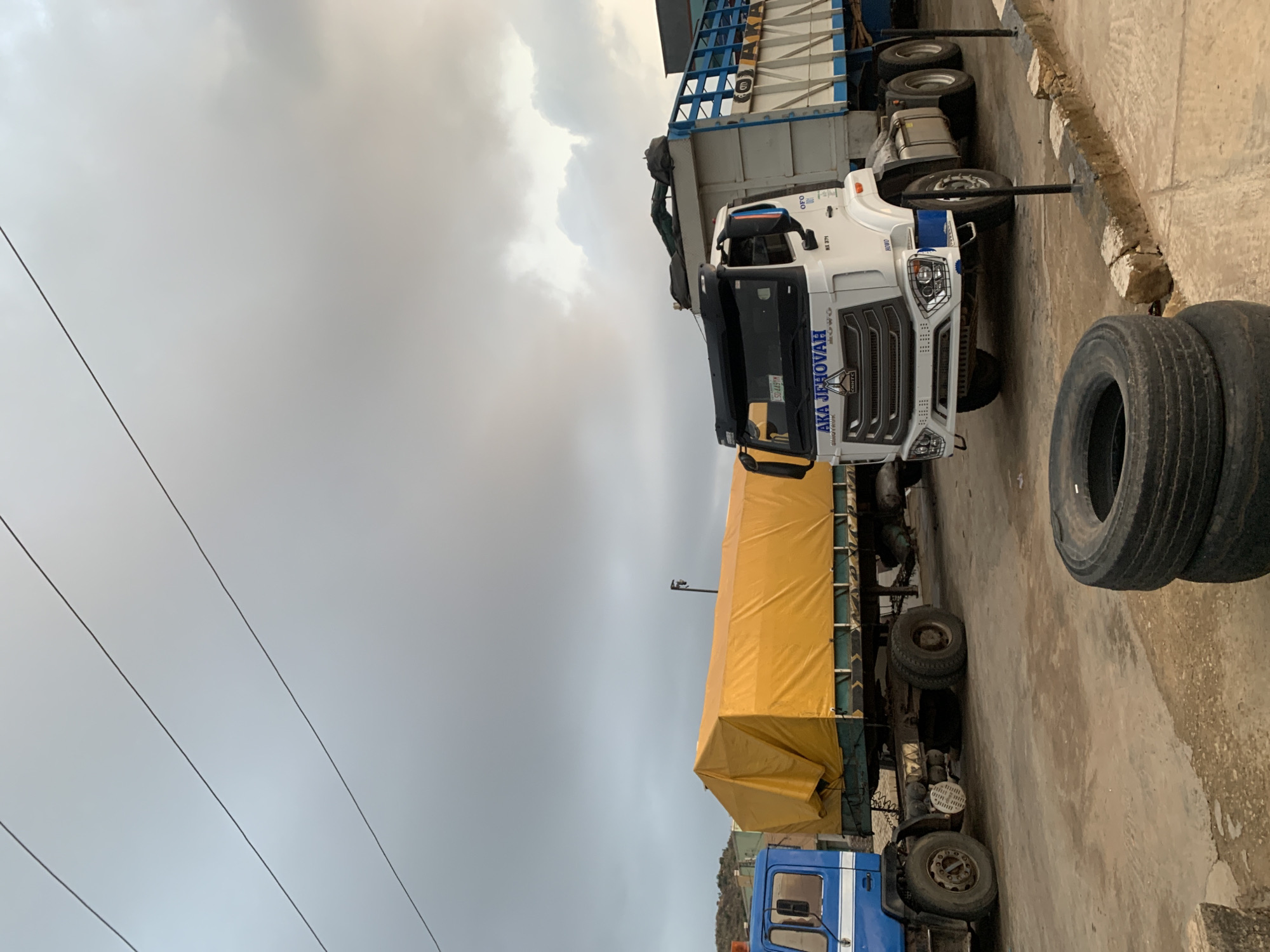 The image shows a parked truck with "AKA JEHOVAH" printed on its cab in Gura Top, Nigeria. The truck has a yellow tarp covering its cargo. Beside the truck, two spare tires are stacked. Overhead, power lines are visible against a cloudy sky. The ground is paved and shows signs of wear.