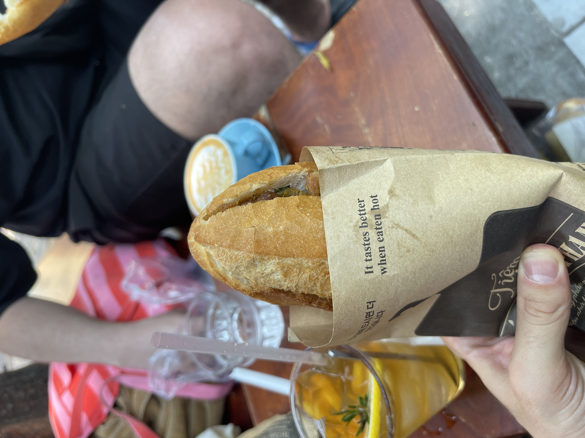 The image shows a partially visible person holding a banh mi sandwich in a paper bag. The sandwich is partly visible, revealing its crusty exterior and fillings. The paper bag has text on it that says "It tastes better when eaten hot," along with a business name in Vietnamese. In the foreground is a glass of yellow liquid with lemon slices and herbs, likely a beverage to accompany the meal.
The scene is set on a wooden table outdoors, which appears to be at a restaurant or cafe. Other items on the table include plastic cups and a drinking straw. In the background, there is a partially visible person wearing dark-colored shorts, and another person wearing a red and pink striped shirt.
The overall setting suggests a casual dining experience in a public space. While Nha Trang, Vietnam, is not discernible from the image itself, it can be stated that is the location.