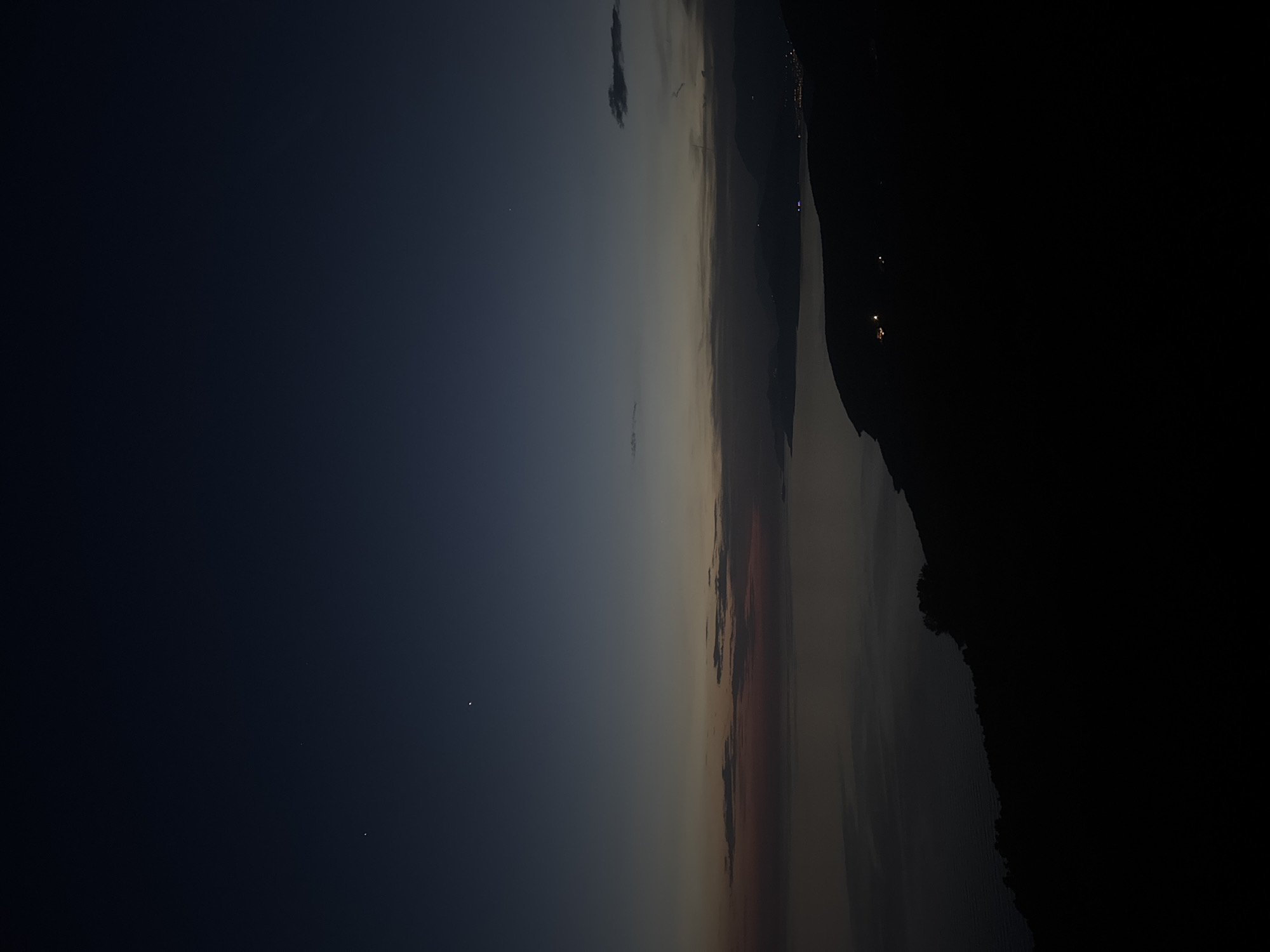 The image captures a landscape at dusk or night in Kovačko Polje, Montenegro. The left portion of the image reveals a dark blue sky with faint stars visible. Moving right, the sky transitions into a lighter gradient of colors, possibly indicating the afterglow of sunset. On the right edge of the frame, a dark silhouette of a mountain or elevated terrain dominates. Faint lights, possibly from buildings or vehicles, punctuate the darkness along the base of the mountain. The orientation suggests a broad view over a valley or plain towards the mountain.