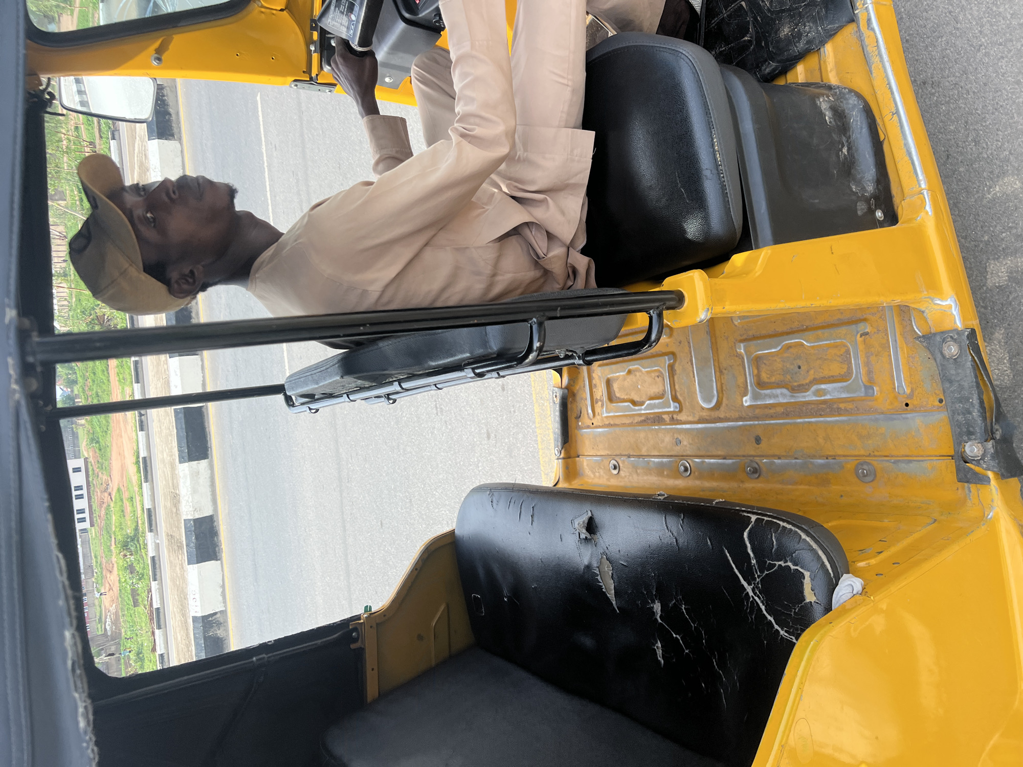 A man is driving a yellow auto rickshaw, possibly in Howayi, Nigeria, based on the assumption of the location. The driver is wearing a beige shirt, pants, and a baseball cap. The interior of the auto rickshaw shows worn black seats. The scene suggests the vehicle is on a paved road with a roadside curb visible through the open side of the vehicle.
