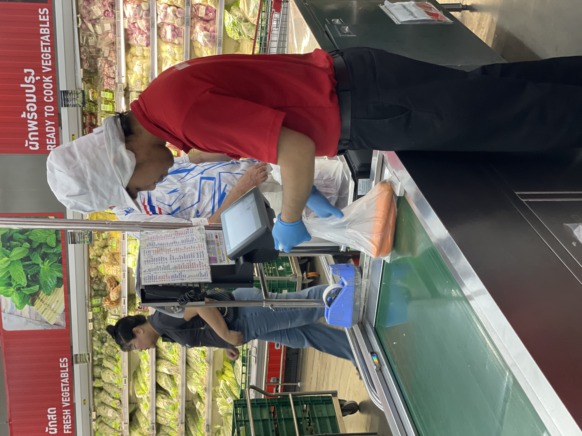 A side profile of a man wearing a red polo shirt, black pants, a white chef's hat, and blue gloves is shown in the foreground of a supermarket in Pattaya City, Thailand. He is standing at a checkout counter, placing a plastic bag of carrots onto a conveyor belt. A woman in a black shirt and blue jeans is visible in the background to the left, walking away from the counter. The supermarket shelves in the background are stocked with various vegetables. The scene appears to be a typical retail environment.