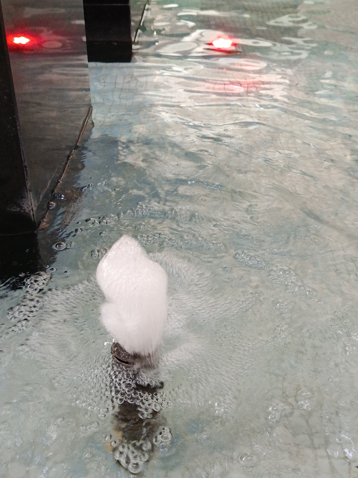 The image depicts a close-up of a water fountain. A jet of water shoots upwards from a dark-colored nozzle, creating a frothy, white effect. The surrounding water is clear, with visible bubbles and ripples radiating outwards from the fountain's base. In the background, there is a blurred section of what appears to be a tiled surface covered in water, with two indistinct red lights reflecting on the surface. The scene is devoid of people, focusing solely on the fountain element and its immediate environment. The location is unknown as Moscow is not evident in the image.