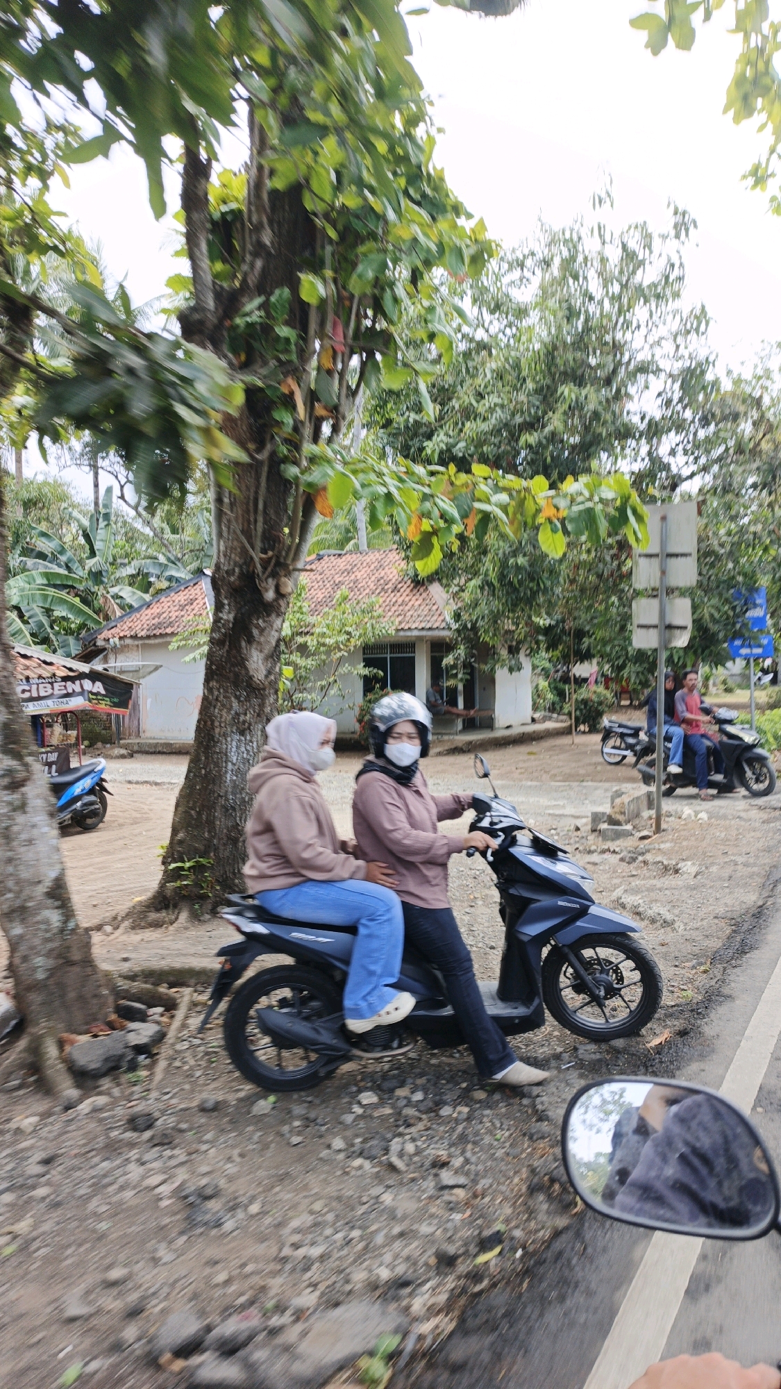 Two individuals are seated on a black scooter. The person in front, wearing a pink hijab and a pink jacket, has a white mask and a helmet. The person behind, wearing a light brown hijab and a light brown hoodie, has blue jeans and light-colored shoes. The scooter is positioned on a dirt and gravel road next to a paved road. In the background, there is a white building with a terracotta roof, surrounded by trees and vegetation. A blue scooter is parked to the left of the building, and another motorcycle with two people is visible in the distance. The scene is set in Cibenda, Indonesia.