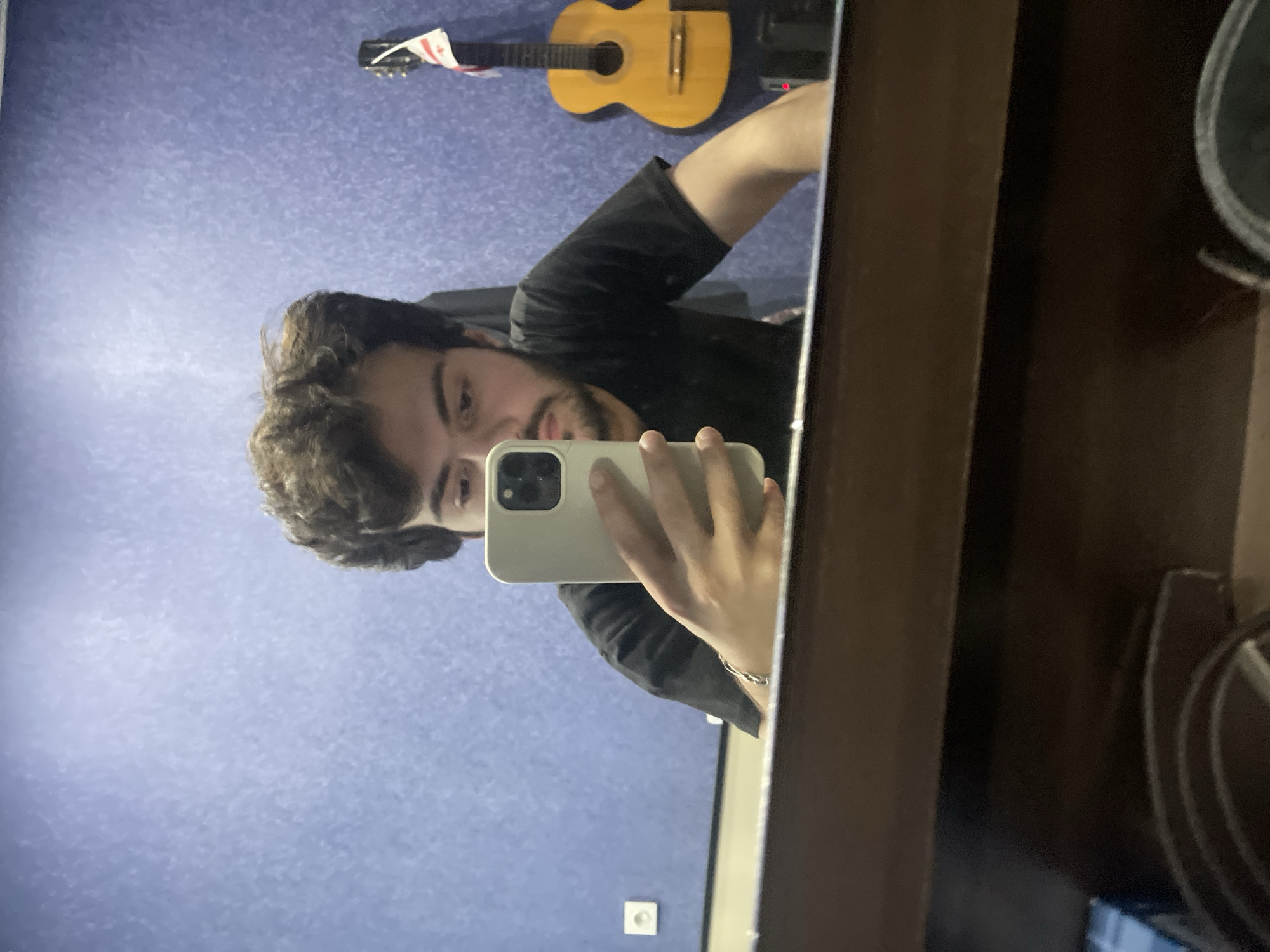 A young man with dark, curly hair and a beard is taking a mirror selfie in a room. He is wearing a black t-shirt and a silver bracelet on his left wrist. He is holding a smartphone with a light-colored case in his right hand. The background is a blue textured wall. To the right of the man, a yellow acoustic guitar with a white flag or banner is hanging on the wall. There is also a white electrical outlet on the wall to the left of the man. The bottom portion of the image shows the reflection of the top of a wooden dresser or cabinet, with what appears to be some dark objects on top of it, possibly a belt or a coil of wires. The scene is not identifiable as being in Tbilisi, Georgia.