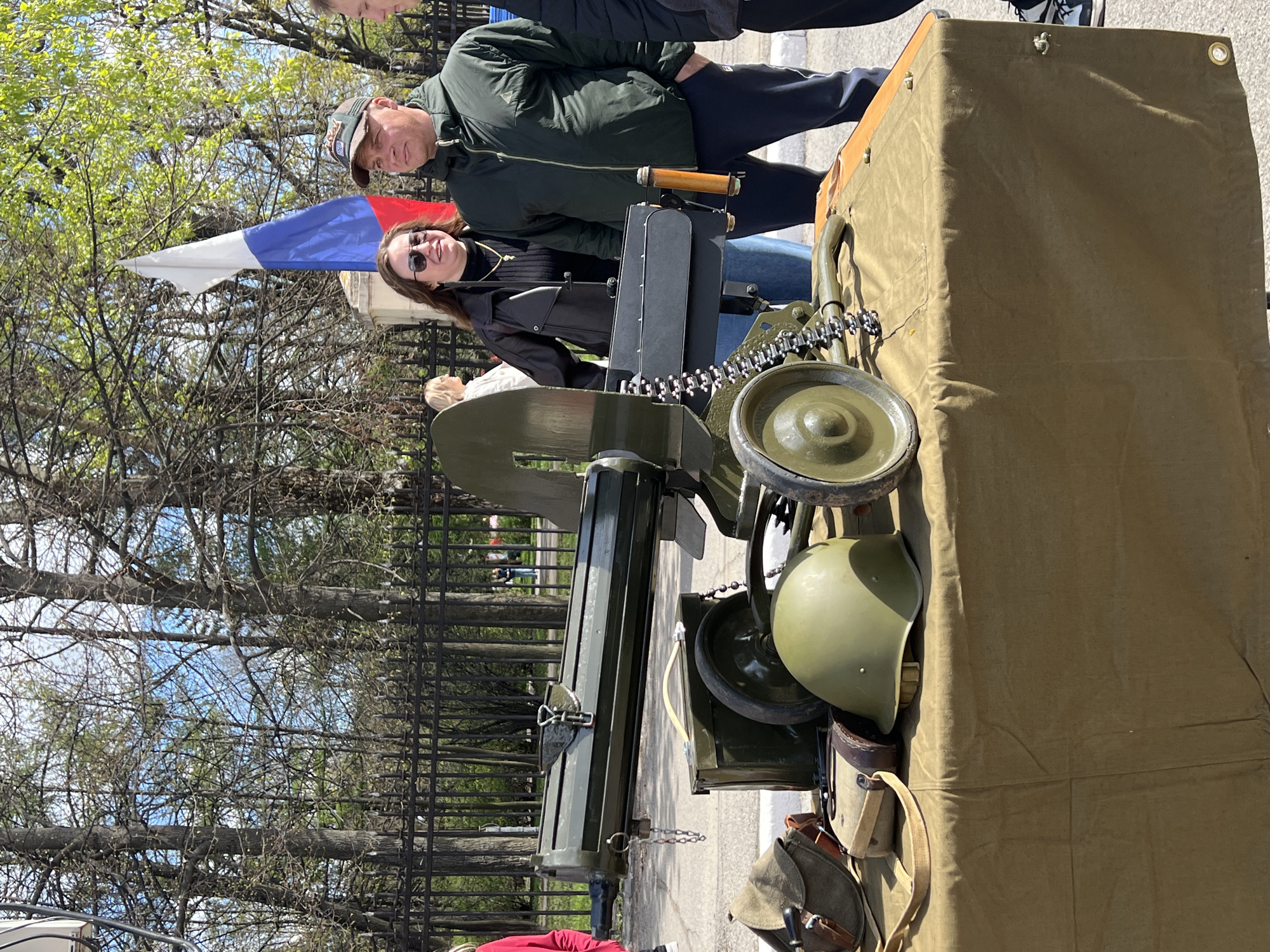 The image shows a military exhibit set up outdoors, possibly as part of a historical display or event. In the foreground, there is a Maxim machine gun mounted on a wheeled carriage, painted in olive drab. A military helmet and canvas pouches are attached to the side of the carriage. The background is blurred. A person wearing sunglasses stands behind the machine gun, and an older man in a cap stands next to them. There is a Russian flag. The presence of a metal fence, trees, and a paved surface indicates an urban park setting. A location in the city of Cherepovets, Russia is not evident based on this image.