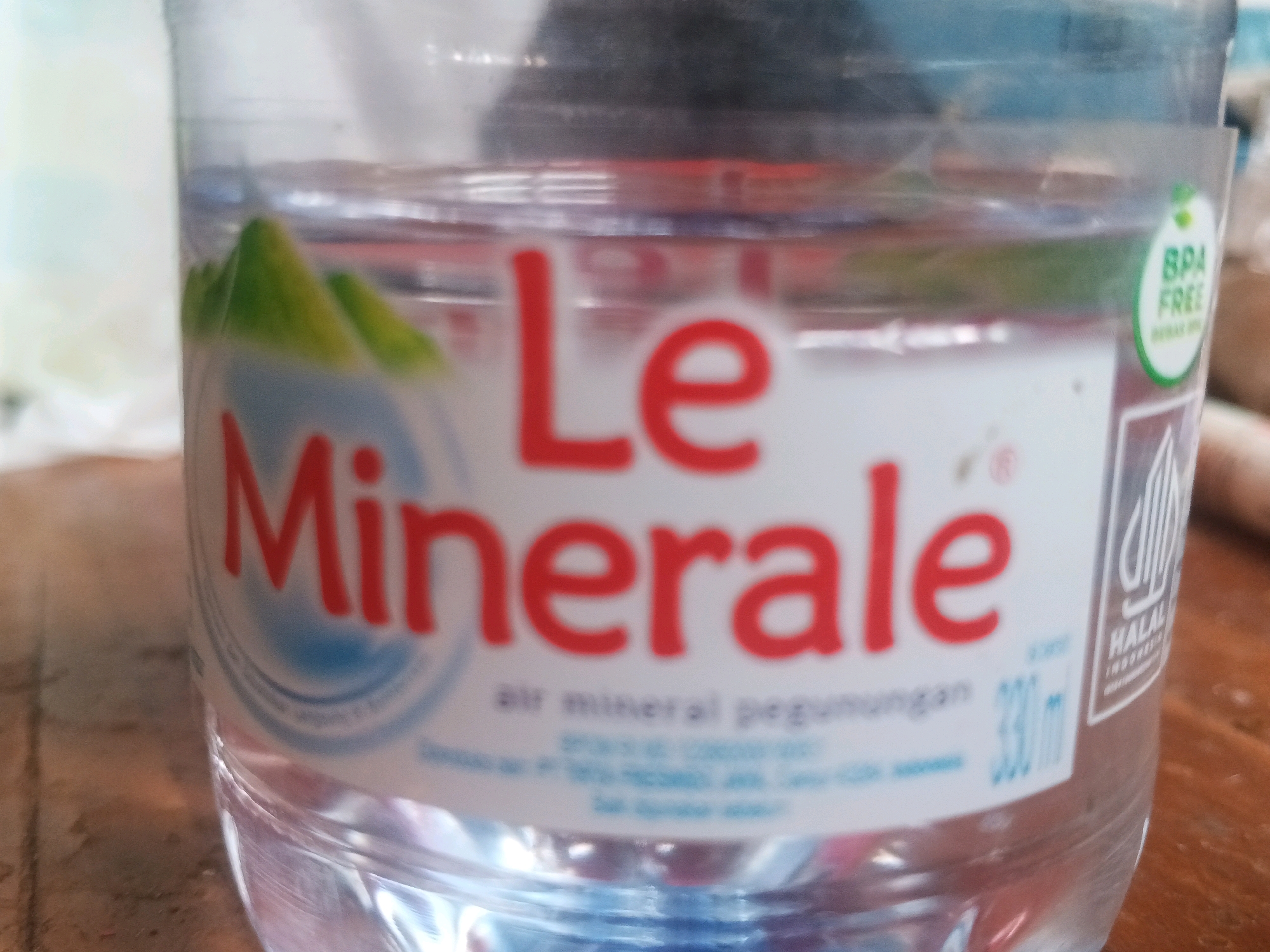 A close-up shot shows a plastic bottle of Le Minerale bottled water. The label is white with red lettering and features an image of green mountains. The label also includes a "BPA FREE" certification and a Halal logo. The bottle is partially filled with water and is placed on a wooden surface, possibly a table. The background is blurred, suggesting an indoor setting. The image is taken from eye level, with the bottle filling most of the frame. The lighting is natural, and there are no people visible in the image. The water is clear and appears to be pure. The bottle's plastic material is transparent.