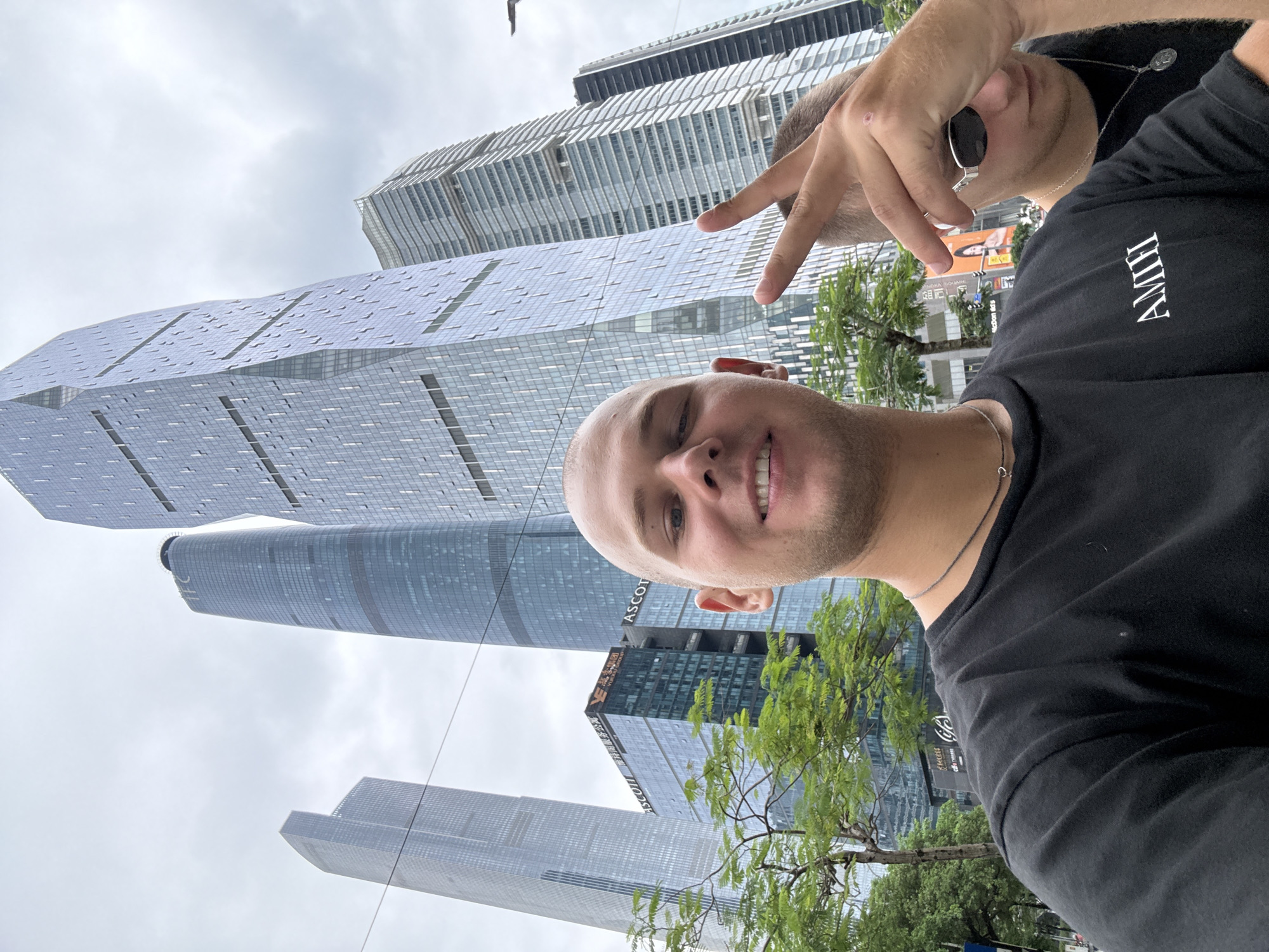 In a selfie taken in Tianhe District, China, two young men stand before a backdrop of towering, modern skyscrapers. The man in the foreground, with a shaved head and a black t-shirt, smiles at the camera, his left hand raised in a peace sign. He wears a simple necklace. To his right, partially obscured and in the background, another young man, wearing sunglasses and a black shirt, looks down. The buildings behind them are architectural marvels of glass and steel, reflecting the overcast sky. Green trees and streetlights are visible in the mid-ground, suggesting an urban park or avenue setting. The overall scene conveys a sense of youthful adventure and appreciation for the modern cityscape.