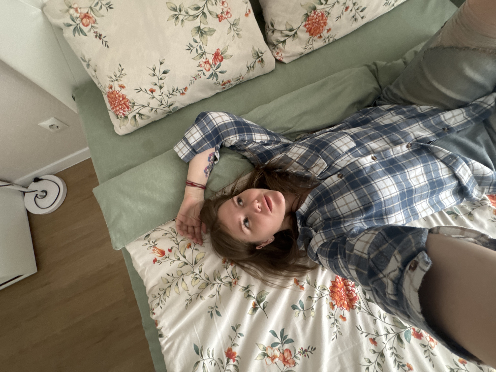 A woman is lying on a bed, taking a selfie. The bed has floral patterned pillows and bedsheets. The woman is wearing a blue and white plaid shirt and jeans. The room appears to be indoors. There is no identifiable location visible in the image, so it is not possible to confirm if it is in Perm, Russia.