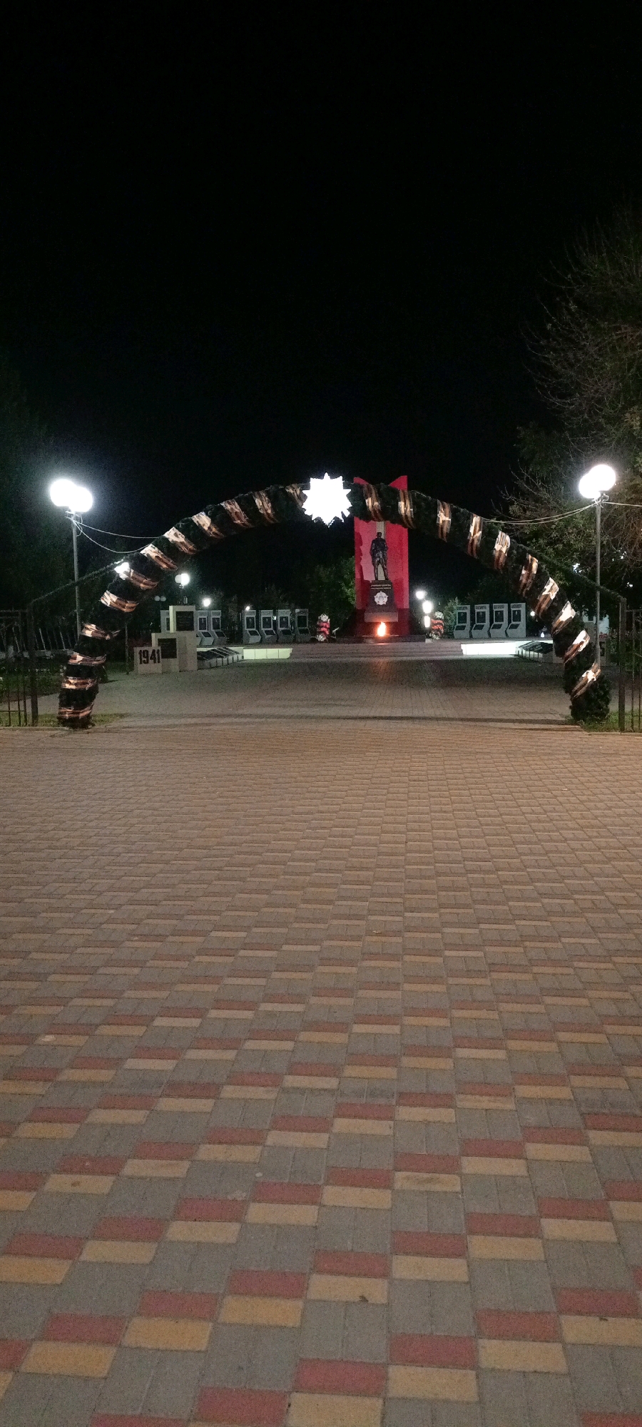 The image shows a night view of a memorial complex in Kirsanov, Russia. In the foreground, there is a paved walkway made of interlocking bricks in shades of red, yellow, and gray. An arch decorated with evergreen boughs and what appear to be memorial ribbons spans the walkway. Beyond the arch, a monument is visible. This monument features a red obelisk with a statue of a soldier on top. A flame burns at the base of the obelisk, indicating a perpetual flame or eternal fire. To the left of the main monument, there are several smaller stone structures that appear to be part of the memorial, including one with "1941" inscribed on it. In the background, there are trees and several streetlights illuminating the scene. The overall atmosphere is solemn and commemorative.
