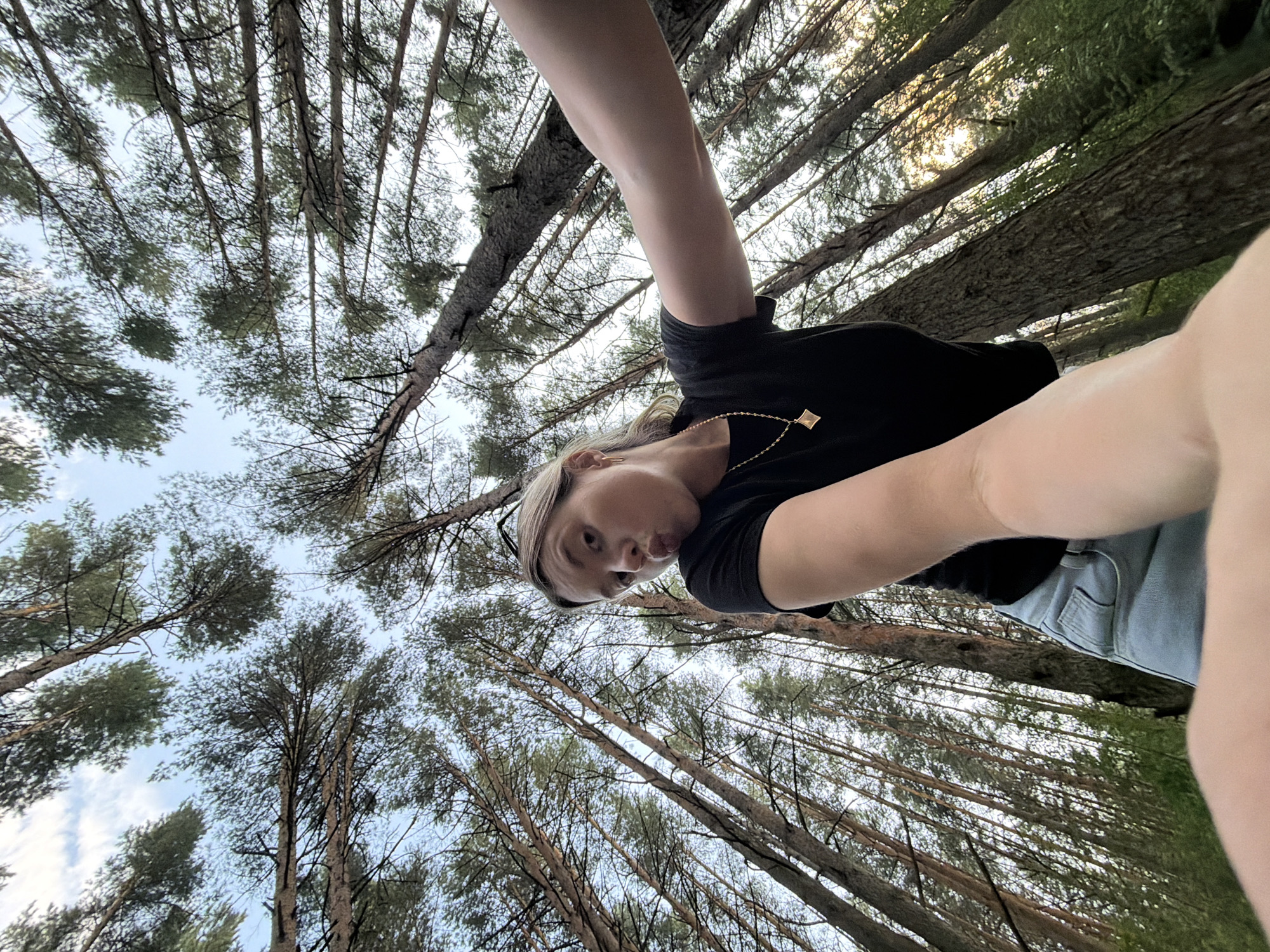 The image is a low-angle, wide-shot selfie taken outdoors in a forest, likely near the city of Chaikovsky, Russia. The foreground features a young woman with blonde hair pulled back, wearing a black t-shirt and light blue denim shorts, and a gold necklace. She is making a puckering face towards the camera, with her right arm extended to take the photo. The background consists of a dense forest of tall, thin pine trees, their trunks reaching upwards towards a partially cloudy sky. The lighting suggests it is either early morning or late afternoon, with sunlight filtering through the trees. The perspective looking up at the trees and the woman creates a sense of immersion in the natural environment.