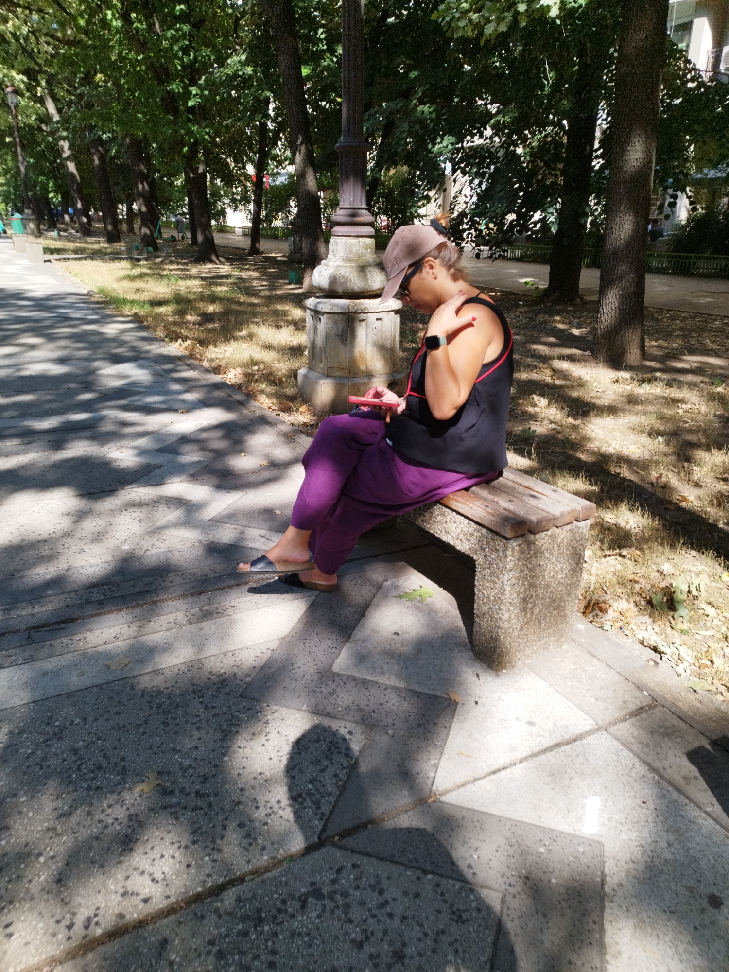 The image depicts a woman sitting on a park bench in Bucharest, Romania. She is wearing a brown baseball cap, sunglasses, a black sleeveless top with red trim, and loose-fitting purple pants. She is holding a pink object, possibly a phone or book, in her hands and appears to be looking down at it. A smartwatch is visible on her left wrist. The bench has a stone base and a wooden seat. The park setting features mature trees casting dappled shadows on a paved walkway and stairs. The overall lighting suggests it is daytime, likely during the warmer months.