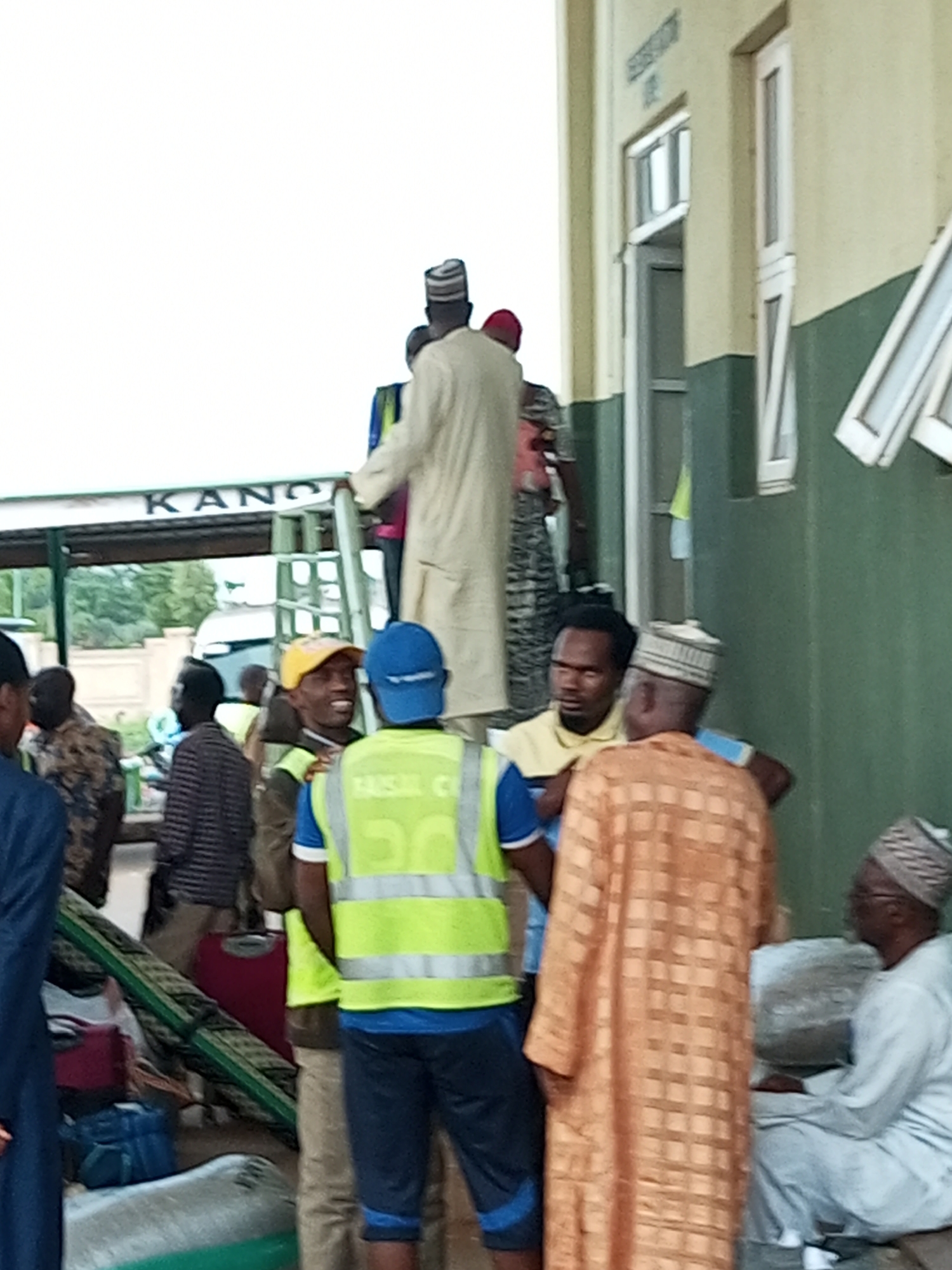 The image depicts a group of people gathered outdoors, possibly at a transportation hub. In the background, a building with a green and beige facade is visible, featuring windows and what appears to be signage. The word "KANO" is partially visible on a green awning. Several individuals are standing and interacting, some wearing traditional attire and headwear. One person is wearing a bright yellow reflective vest. Luggage is scattered on the ground. The overall scene suggests a bustling environment with people preparing for travel or having just arrived. The context points towards the city of Gombe, Nigeria, likely a bus station or similar transit point given the presence of luggage and the informal gathering.