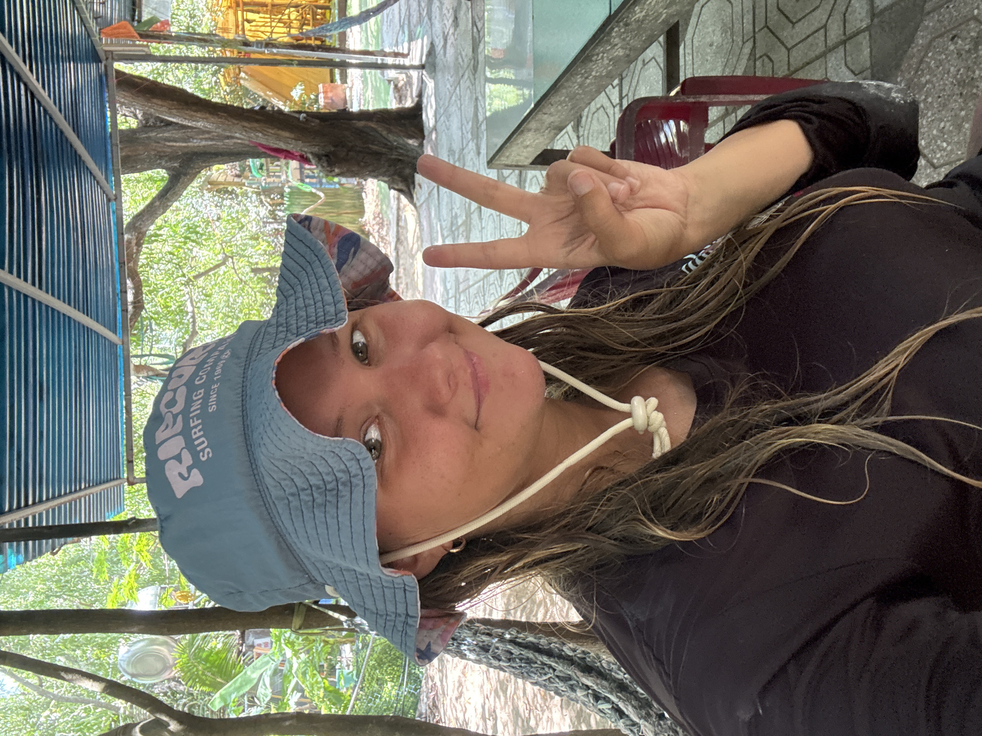 In an outdoor covered area with a slatted blue roof and visible trees and foliage, a young woman is taking a selfie. She is wearing a light blue bucket hat with white lettering that reads "RIPCURL SURFING COMPANY SINCE 1969". She has long, wet, light brown hair, a dark long-sleeved shirt, and a white rope tied at the neck. She is holding up her right hand in a peace sign gesture. Behind her, a hammock is visible to the left, and a red chair and a table with a reflective surface are visible to the right. The ground is tiled. The environment appears to be tropical. The location is the city of Cam Lam, Vietnam.
