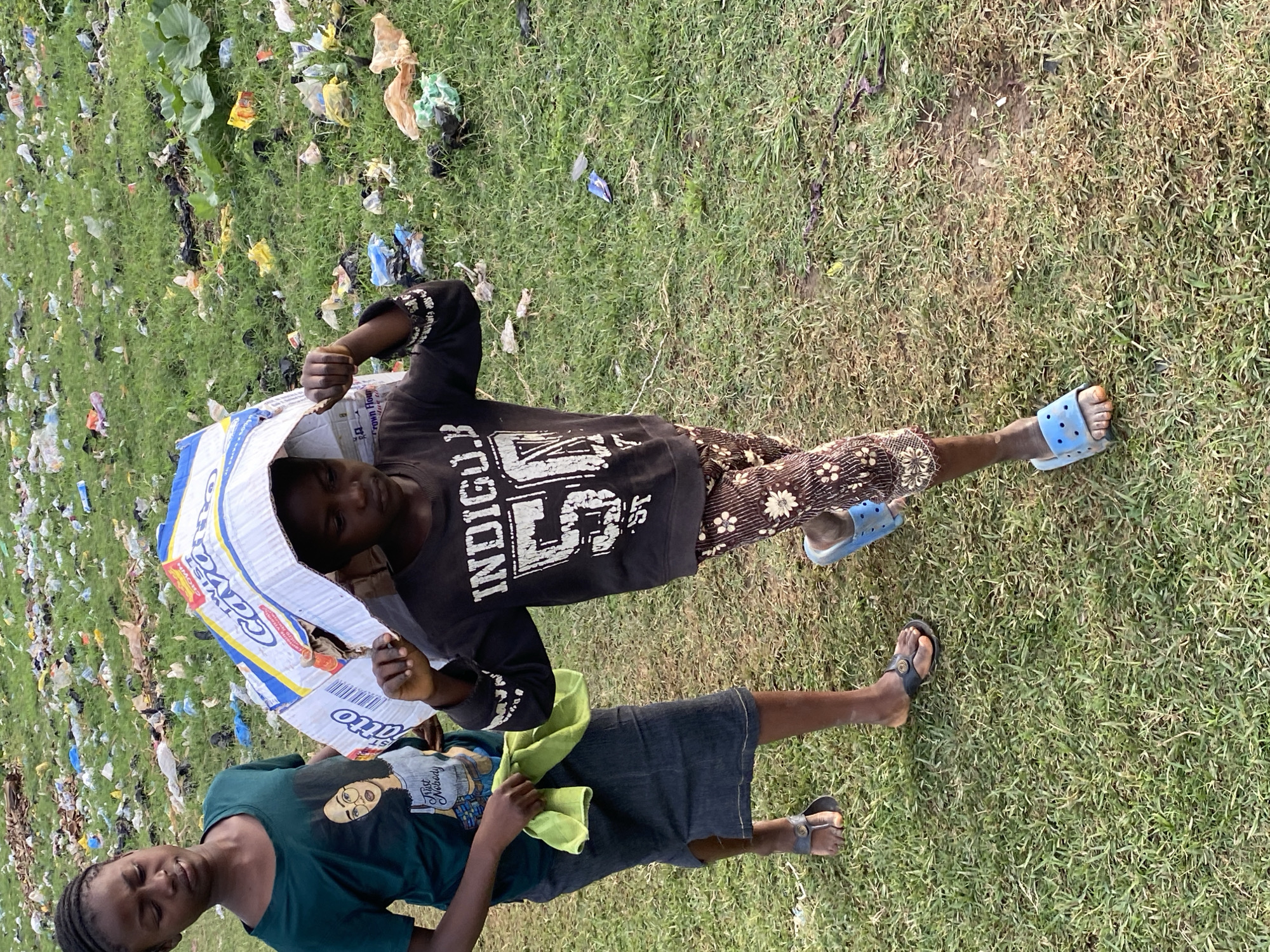 Two young individuals, likely children, are walking across a grassy area littered with trash. The individual on the left, wearing a dark green t-shirt with a graphic of a woman and a dark denim skirt, carries a light green cloth in their left hand. The individual on the right, wearing a dark long-sleeved shirt with "INDIGO" printed across the chest and patterned pants, is carrying a large cardboard box over their head as a makeshift shelter from the sun. The background is filled with a significant amount of plastic waste, garbage bags, and various discarded items scattered across the ground, indicative of an unmanaged waste disposal area. The scene appears to be located outdoors in an urban or semi-urban environment, possibly a field on the outskirts of Bukuru, Nigeria, given the context. The ground is uneven with patches of dry earth and green grass.
