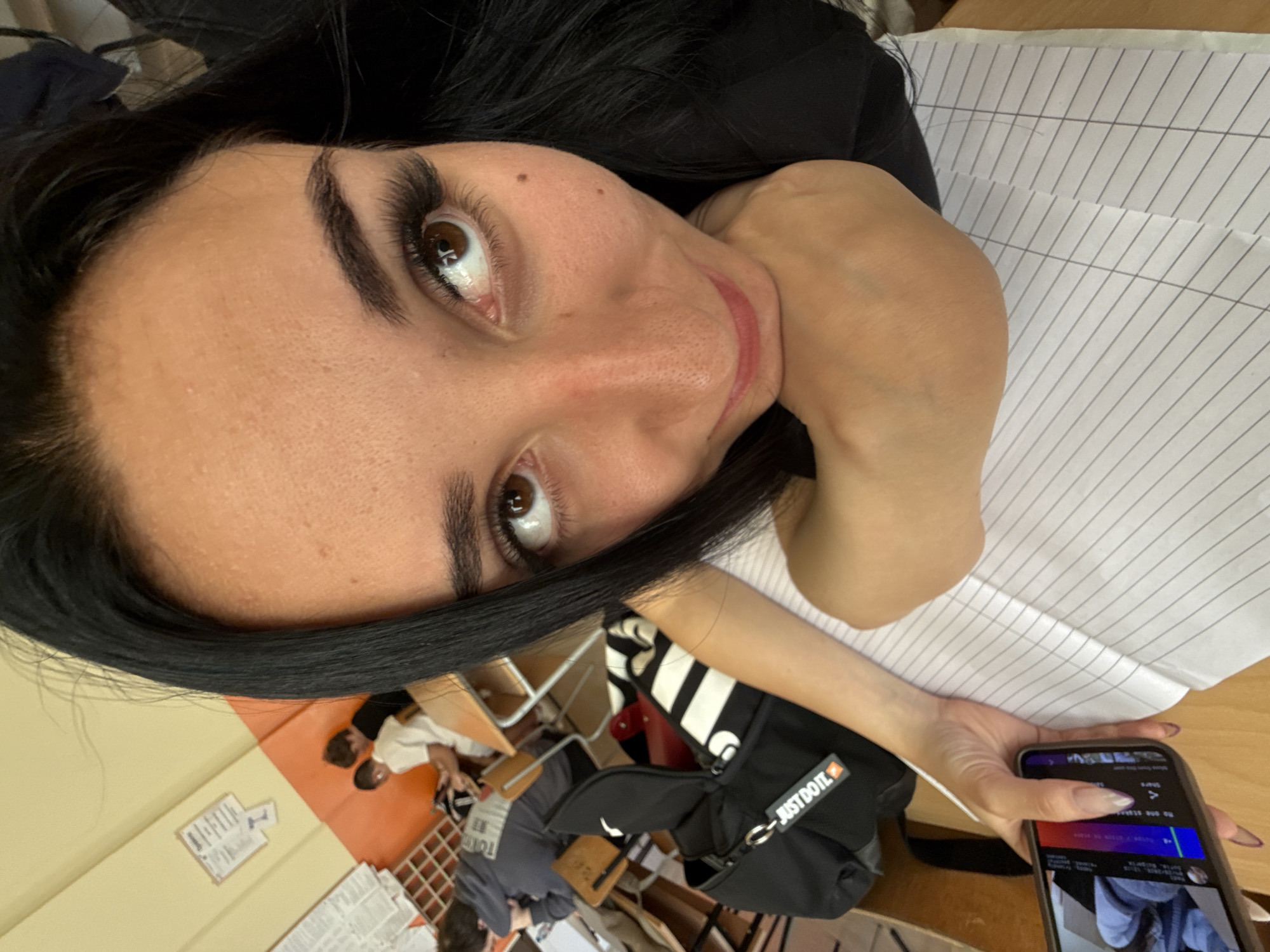 A woman with dark hair is taking a selfie with a cell phone. She is indoors, seemingly in a classroom or office setting. There are desks, chairs, and other people in the background. A black "Just Do It" Nike bag is visible. Lined paper is also present. Sofia, Bulgaria cannot be determined from the image.