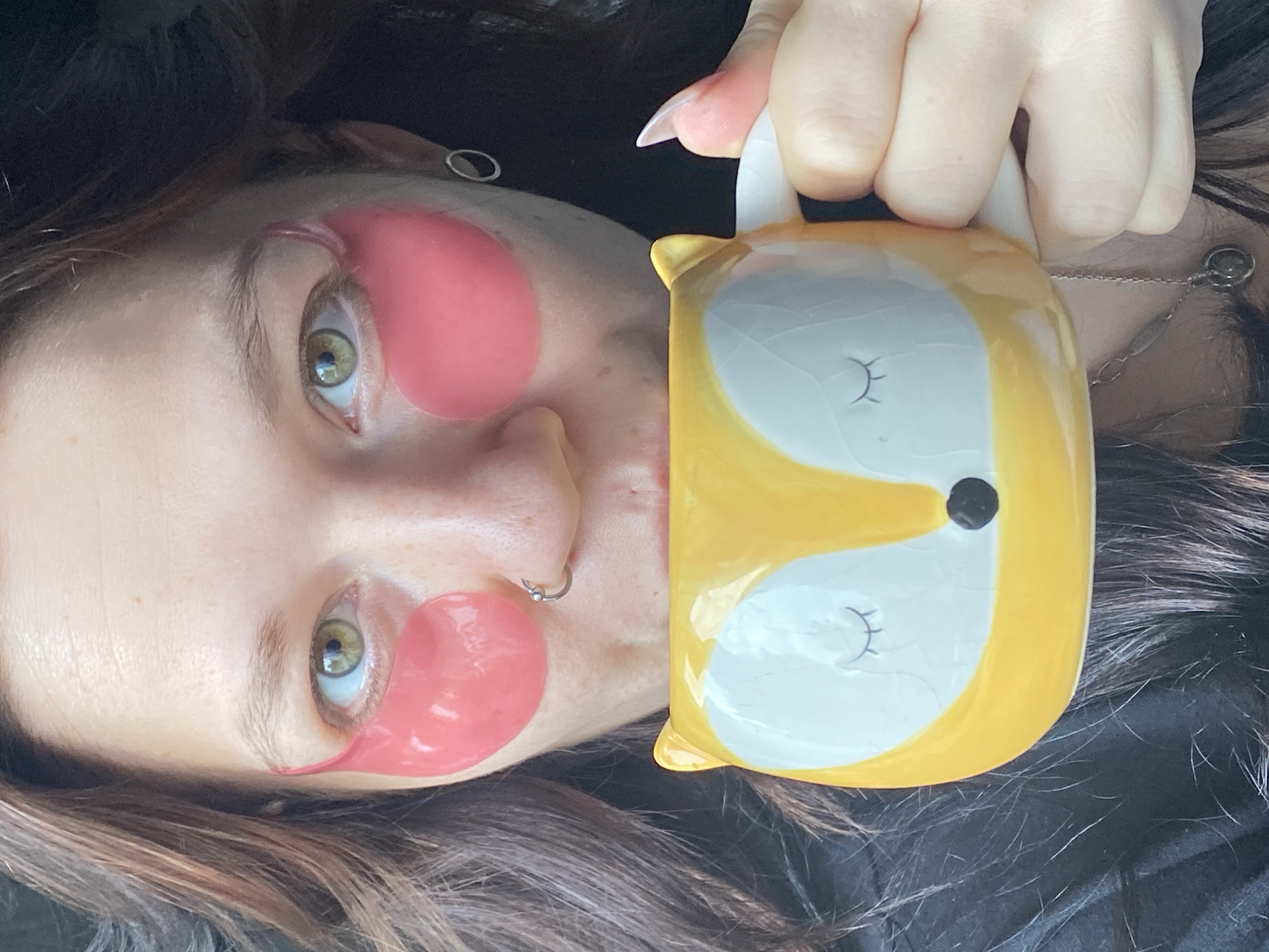 An image, rotated 90 degrees counter-clockwise, depicts a person lying down, engaged in a self-care routine in Chaikovsky, Russia. The individual, likely a woman, has dark brown hair and striking green eyes. They are wearing two vibrant pink, gel-like under-eye patches, one beneath each eye. A silver hoop nose ring is visible on their left nostril, and a simple silver ear cuff adorns their right earlobe.
The person is holding a distinctive yellow and white ceramic mug, designed to resemble a stylized fox face with closed, smiling eyes and a small black nose. The mug is held firmly by a hand with long, well-kept fingernails that appear to have a light-colored polish. A delicate silver chain necklace with a small circular pendant rests on their chest/neck area, partially obscured by their hair. The background is dark and out of focus, suggesting an indoor, comfortable setting. The overall atmosphere is one of relaxation and coziness. No text is visible in the image.