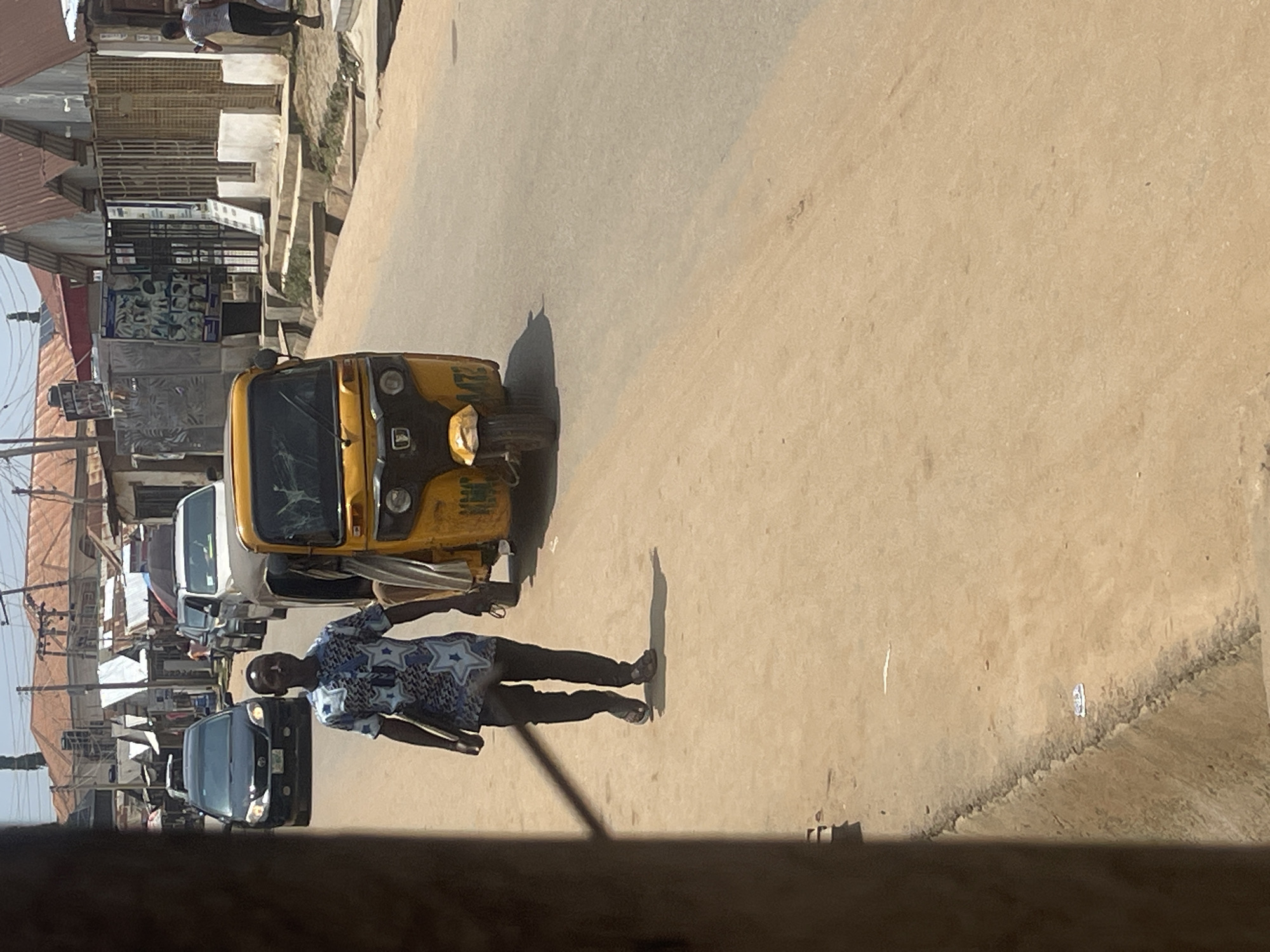 A man in a blue and white patterned shirt and black pants walks on a dusty road in a city. He carries a flat object under his left arm. To his right, a yellow auto-rickshaw is parked, its front wheel turned to the left. Behind the man, a dark-colored sedan is visible on the road, followed by a white van. In the background, buildings with corrugated metal roofs line the street, and power lines stretch across the sky. The scene suggests a typical urban environment in Abuja, Nigeria, with moderate vehicle and pedestrian traffic.