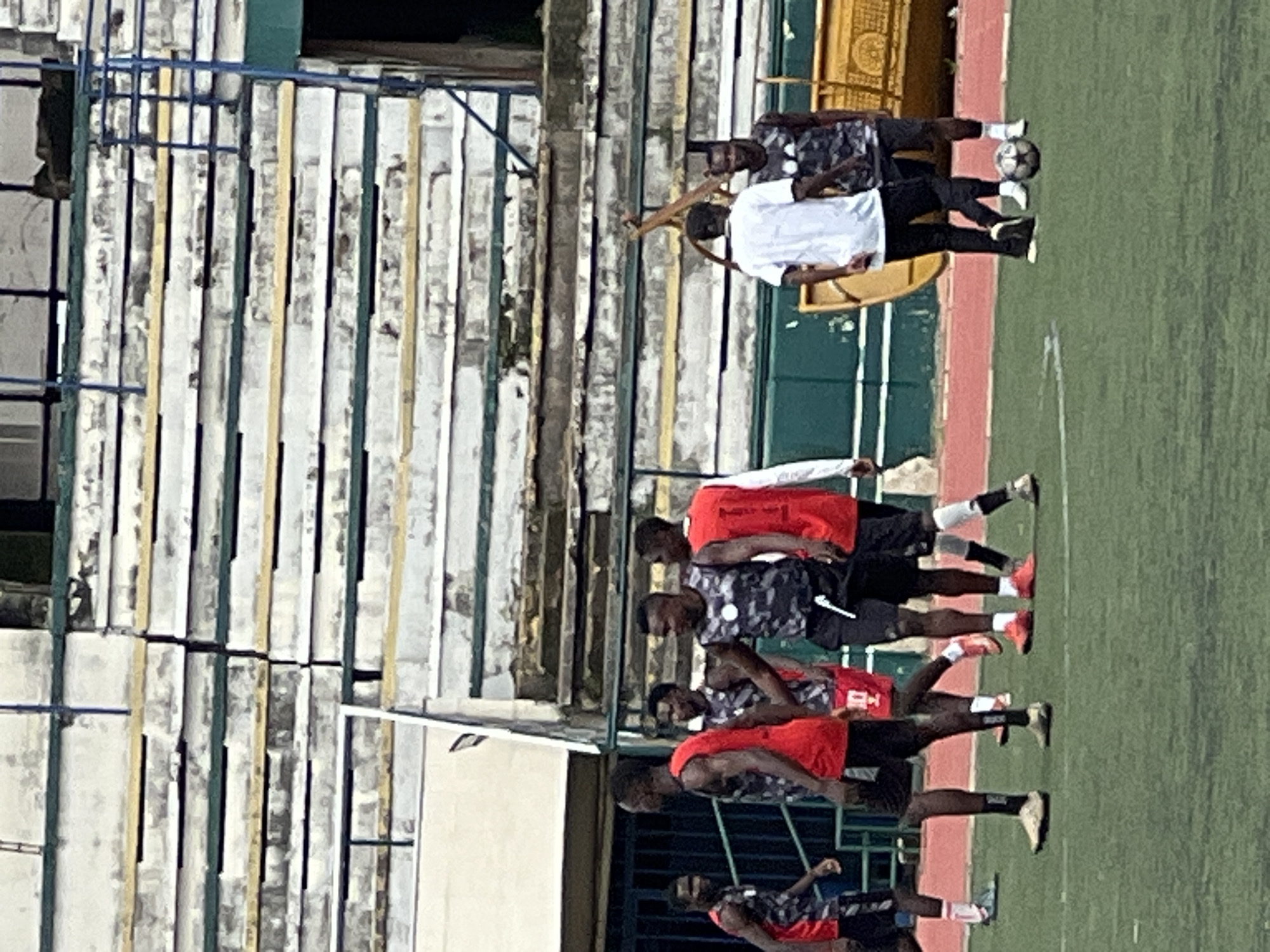 A group of six African men in athletic attire are on a green turf field, likely a soccer pitch, in front of a dilapidated stadium seating area. The stadium appears to be an outdoor structure with weathered, multi-tiered concrete seats and some exposed rebar. In the background, a yellow, open-top vehicle, possibly a truck or a large cart, is visible. One man in a red jersey with the number 11 on the back is standing with his hands on his hips. Another man in a red jersey and black shorts is walking. Two men are wearing camouflage-patterned sleeveless tops and black shorts. One of these men has his arm around the shoulder of the man in the red jersey. Two other men are in the foreground; one is wearing a white t-shirt and black pants, and the other is wearing a camouflage sleeveless top and black shorts. A soccer ball is on the ground near these two men. The lighting suggests it is daytime. The overall scene conveys a sense of a casual sporting gathering or practice. The stadium seating's condition and surrounding environment suggest a location in Rafin Par, Nigeria, which is known for its outdoor recreational facilities and local sporting events.