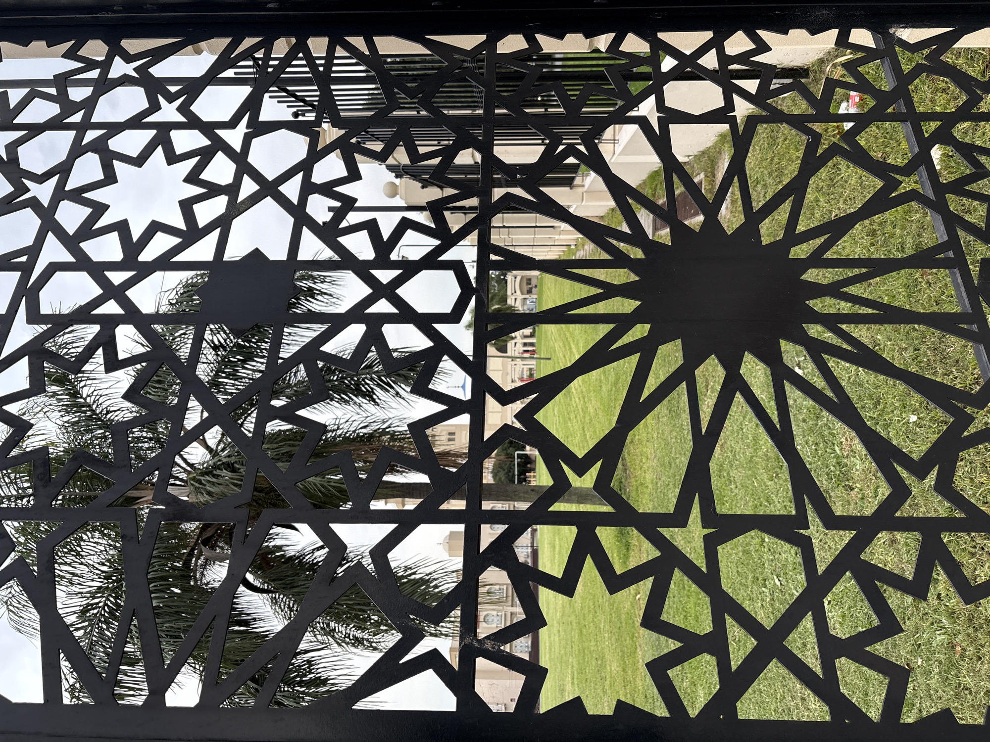 An eye-level, medium shot presents a square section of a dark metal gate adorned with an Islamic star pattern. Through the gate, a landscape view unfolds, with a palm tree visible on the left and a patch of green grass on the right. In the background, a building with architectural details can be seen. The sky is overcast, suggesting a cloudy day. The location appears to be in Buenos Aires, Argentina.
