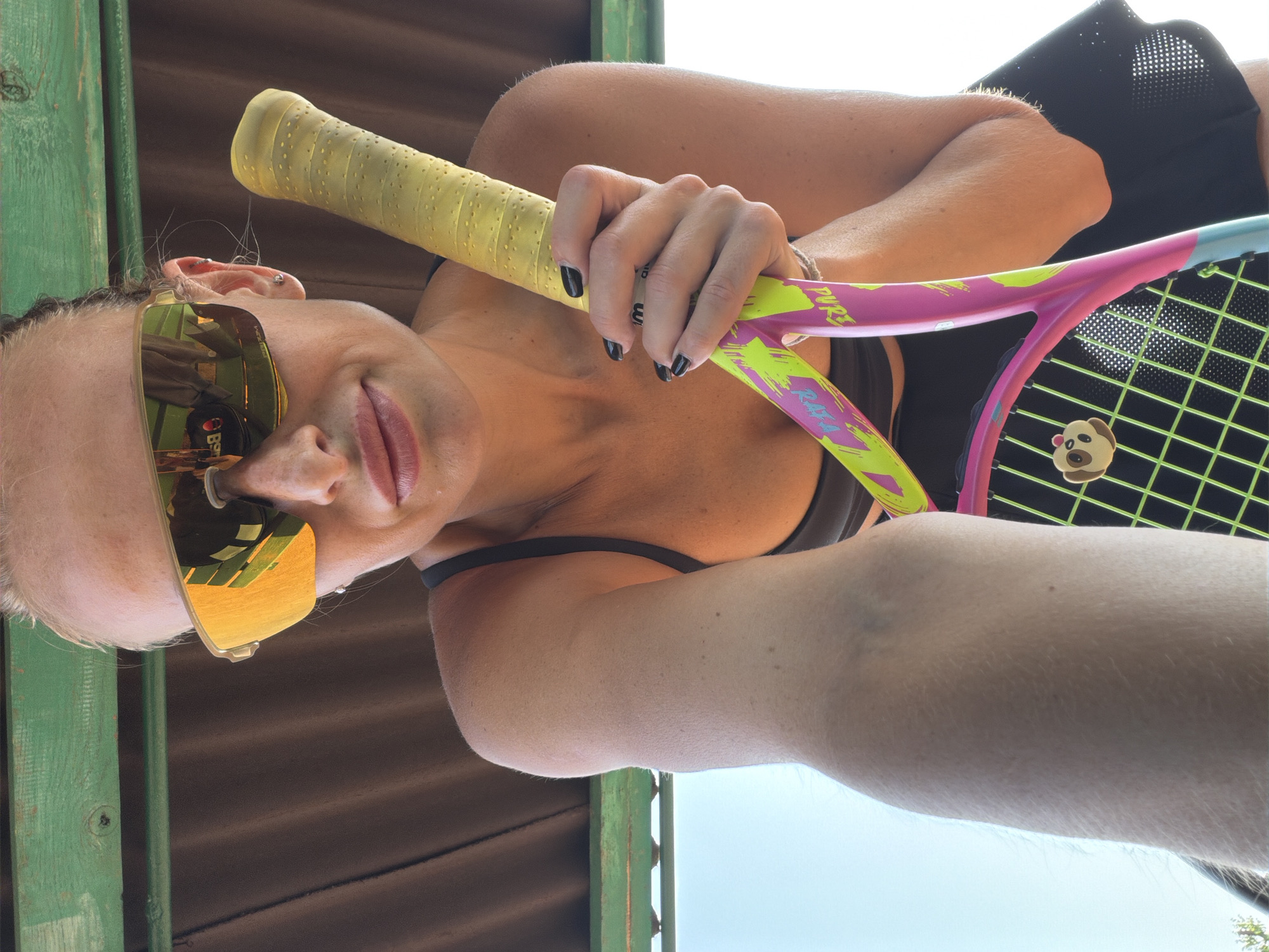 A woman wearing large, yellow-tinted sunglasses and a black sports bra holds a pink and yellow tennis racket. She has blonde hair pulled back and is wearing a black skirt. The background appears to be an outdoor setting with a green-painted wooden structure overhead and a bright blue sky. The tennis racket has a yellow grip and neon green strings. A small, light brown cartoon dog charm is attached to the racket strings. The image is a low-angle selfie. No specific location in Krasnoyarsk, Russia is discernible from the image.