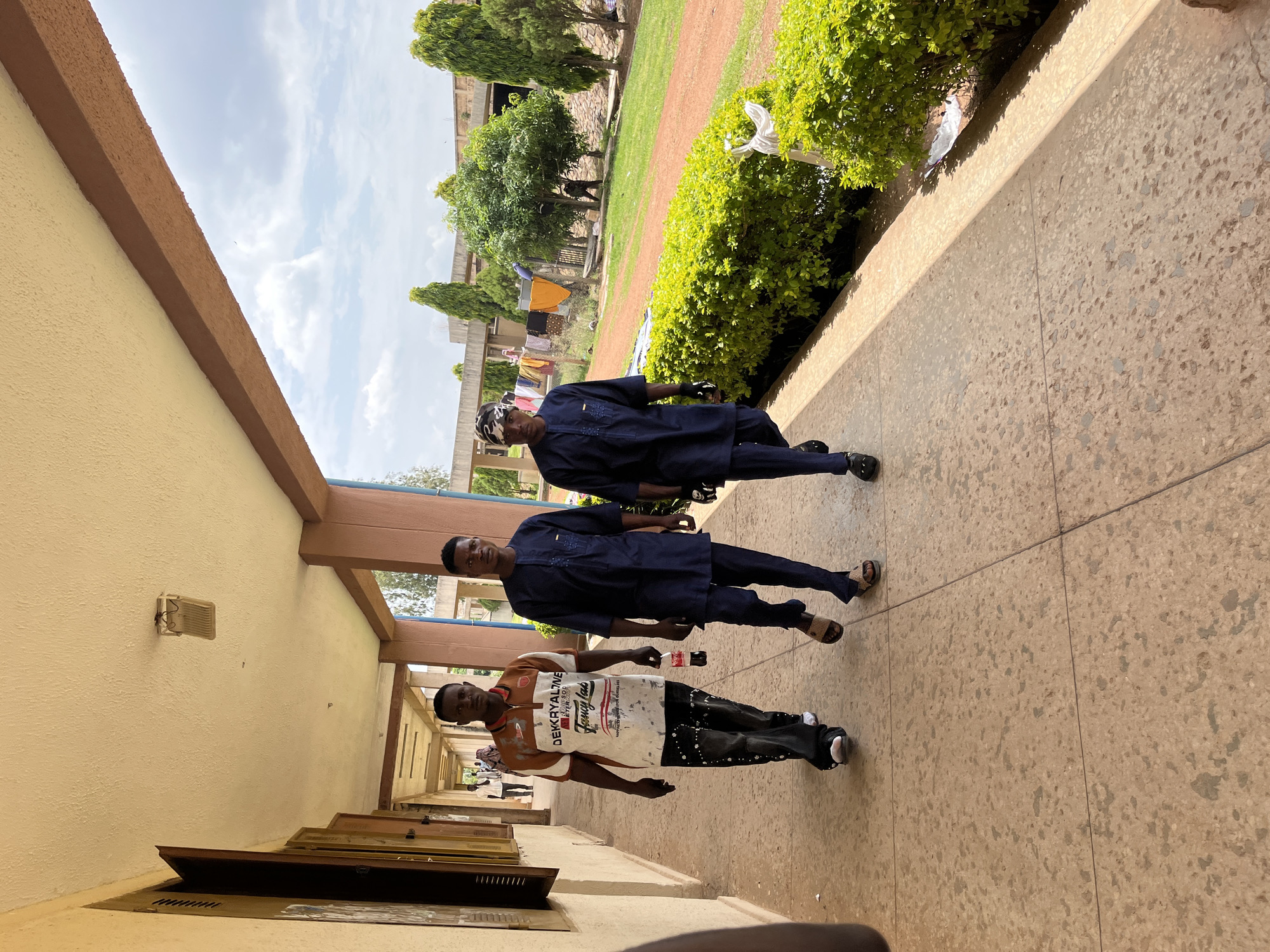 Three men are standing on a sidewalk. Two are wearing dark blue shirts and pants, and one is wearing a white t-shirt and dark pants. The location is Gwong, Nigeria. The sidewalk is adjacent to a building with a beige facade, including window and vent. There is foliage to the right of the sidewalk. Laundry hangs to dry in the background.