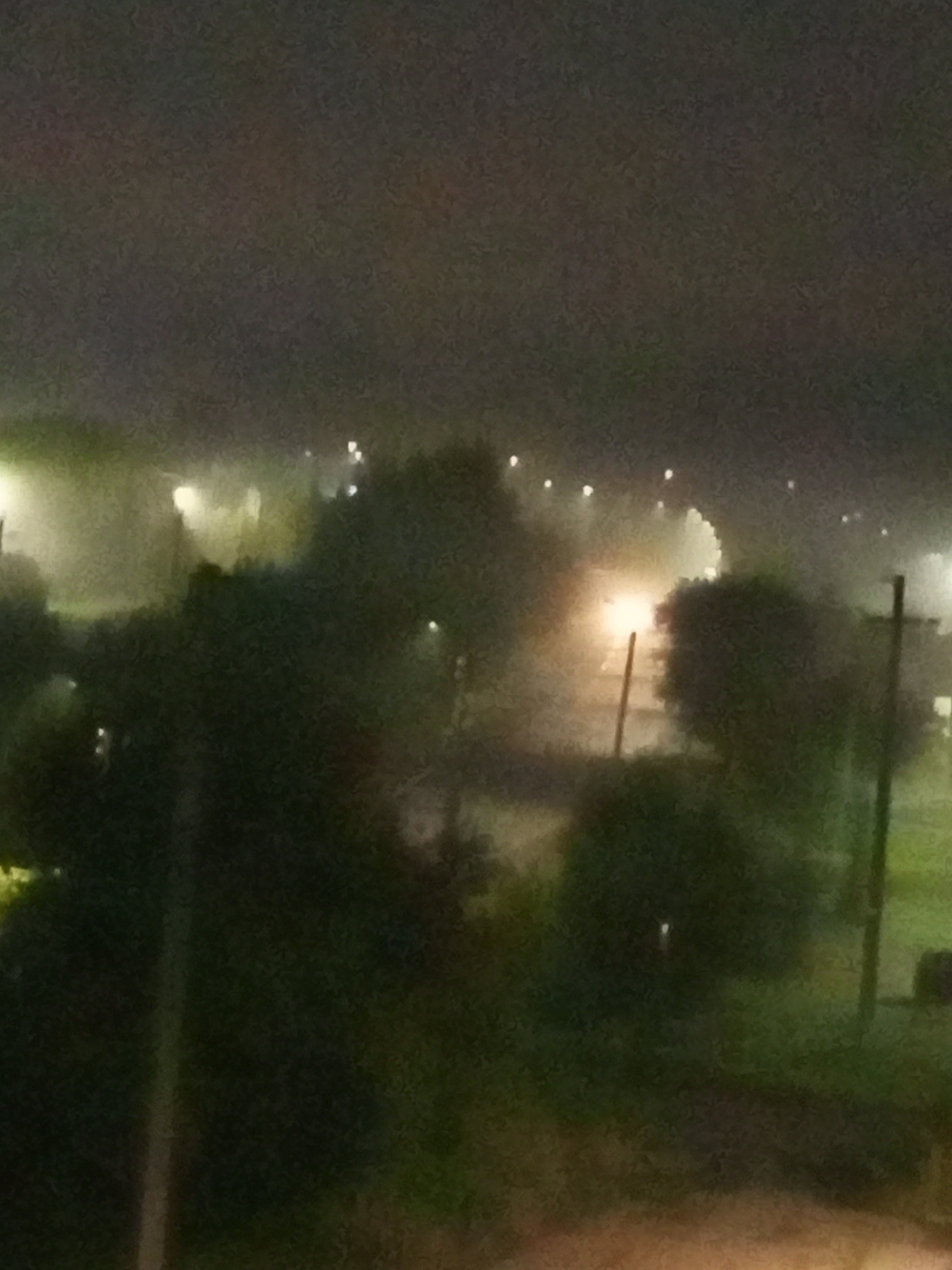 The image depicts a nighttime scene, likely in Chistopol, Russia, characterized by low visibility due to fog or mist. In the foreground, dark, indistinct foliage obscures the view. In the midground, a bright, glowing light source, possibly headlights of a vehicle or a streetlamp, illuminates the fog, creating a diffused glow. This light source appears to be situated behind a thin, vertical pole-like structure, which could be a traffic signal or a signpost. Further in the background, a row of faint, white lights stretches across the horizon, suggesting the presence of buildings or distant streetlights. The overall atmosphere is dark and somewhat hazy, typical of a dimly lit, foggy night in an urban or suburban environment. There are no discernible people or specific identifiable landmarks.