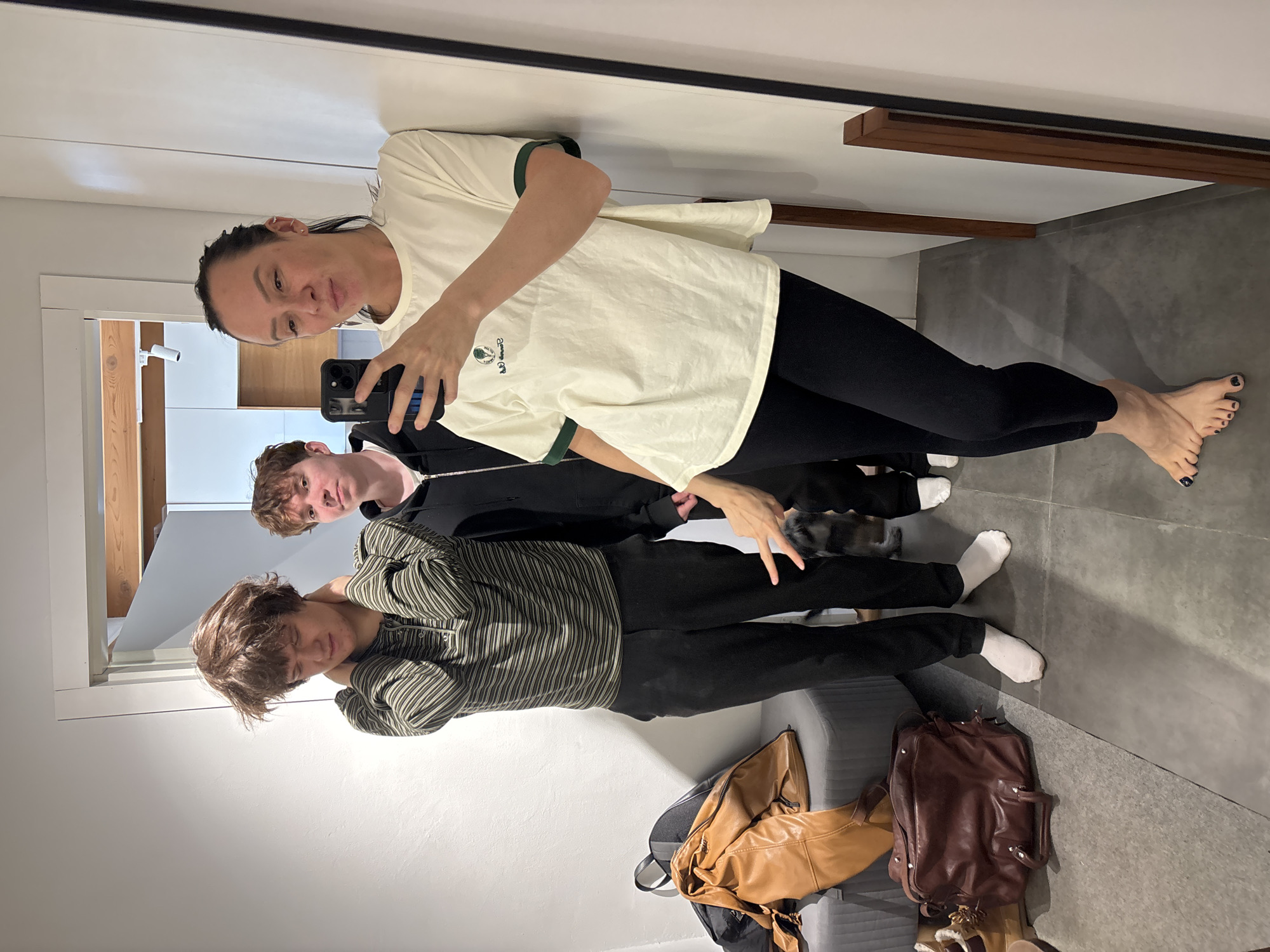 A mirror selfie captures a woman, two younger males, and a small dark-furred pet in an indoor setting. The woman, positioned on the right, holds a smartphone, wearing a cream t-shirt with dark green trim and black leggings. Her dark hair is pulled back, and her toenails are painted a dark color; she is barefoot. A small logo on her t-shirt appears to be "LACOSTE" with an alligator symbol.
To her left, a male with light brown hair wears a dark zip-up jacket, looking towards the camera with a neutral expression. Further left, another male with medium brown hair wears a striped greenish-brown and light-colored long-sleeved shirt, black pants, and white socks, with a relaxed or slightly tired expression. The small pet is partially visible near their legs.
On the lower right, a light brown leather shoulder bag and a darker brown leather backpack/duffel bag rest on what appears to be a bench or seat, with a pair of light-colored furry shoes also visible. The background consists of modern white walls, a grey floor, and wooden architectural accents, including a doorframe and a horizontal beam. The scene is well-lit, suggesting daytime or bright indoor lighting. The photo was taken in Chaikovsky, Russia.