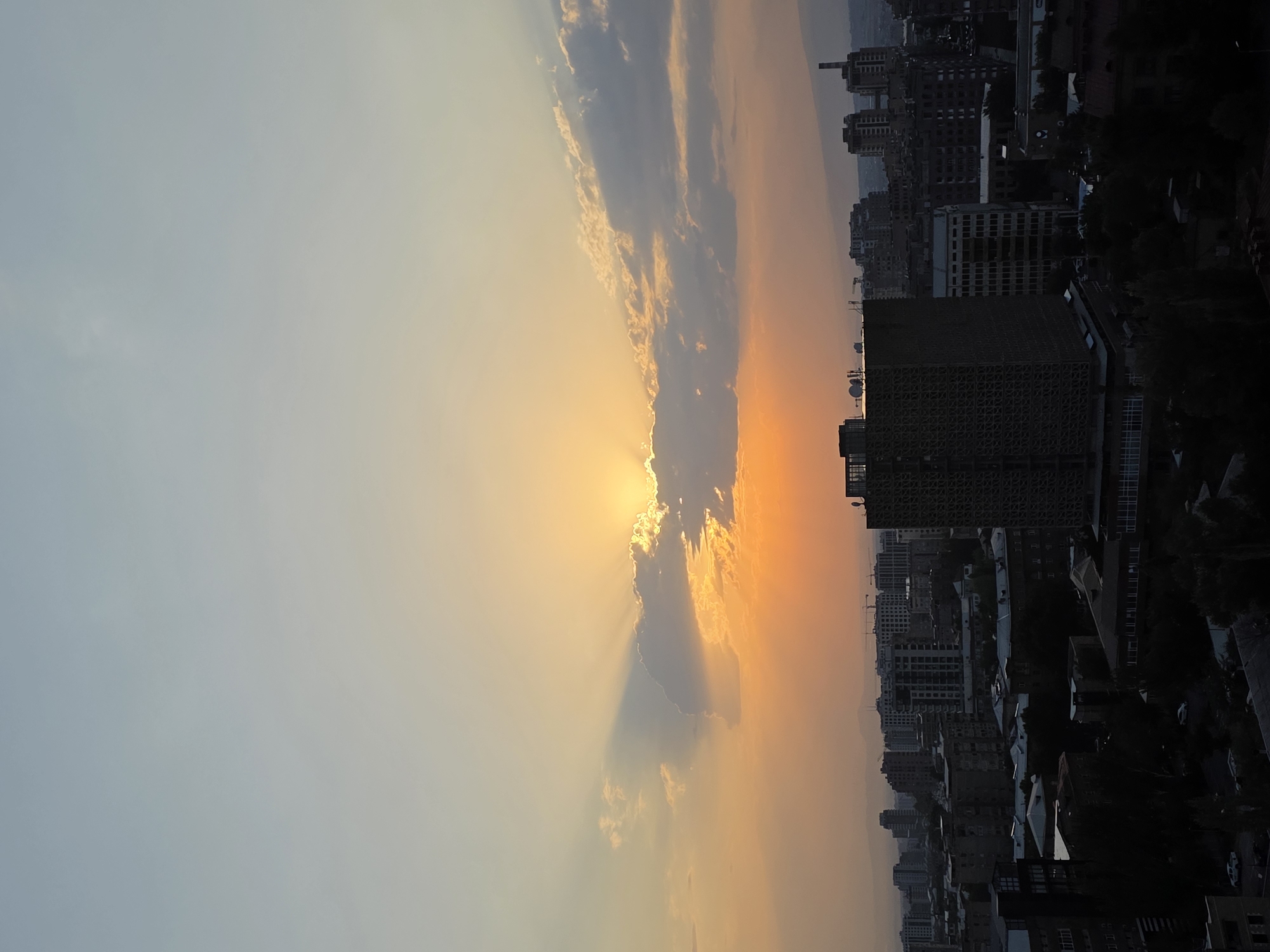 This is a vertical, medium-shot photograph capturing a sunset over Yerevan, Armenia. The sky is dominated by a gradient of colors, transitioning from a pale blue at the top to a vibrant orange and yellow near the horizon. Sunbeams pierce through scattered clouds, creating a dramatic effect. Below the sky, a cityscape unfolds with numerous buildings of varying heights and architectural styles. The foreground features a dark silhouette of trees and rooftops, obscuring much of the lower levels of the buildings. In the middle ground, a prominent, tall, square building with a patterned facade stands out. Beyond this, a dense urban landscape stretches towards the horizon, with more apartment blocks and industrial-looking structures visible. Construction cranes are also spotted amongst the buildings, indicating ongoing development. The overall impression is one of an urban environment bathed in the warm, fading light of sunset.