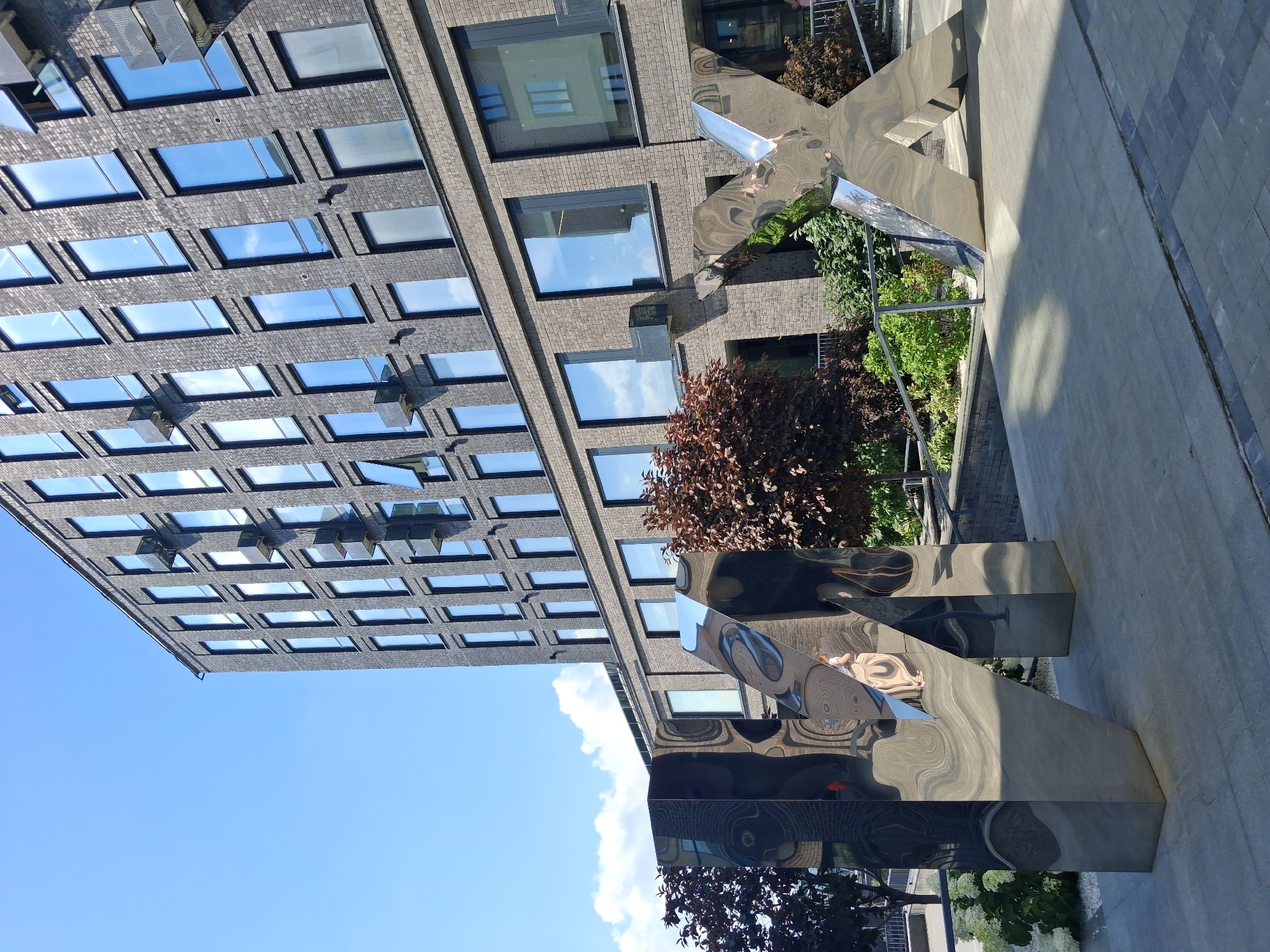 A contemporary brick building with numerous reflective windows dominates the background of this outdoor image. In the foreground, large, polished metal sculptures in the shape of letters "UX" are positioned on a paved surface. The sculpture on the left, representing "U", is a tall, rectangular prism with rounded edges and a highly reflective surface, mirroring the surroundings. To its right, the sculpture representing "X" is also made of polished metal, with two angled triangular forms creating the letter's characteristic shape. Behind the sculptures, a lush green garden area with dark-leaved trees and white flowering bushes is visible. A person, partially obscured, stands behind the "X" sculpture, facing away from the camera. The scene appears to be a public space in front of an office or residential building. The weather is clear with a bright blue sky and scattered clouds. The building's architecture suggests a modern urban setting. This scene is located in Novosibirsk, Russia, specifically in the Novosibirsk State University campus, near the Laboratory of Intellectual Systems building.