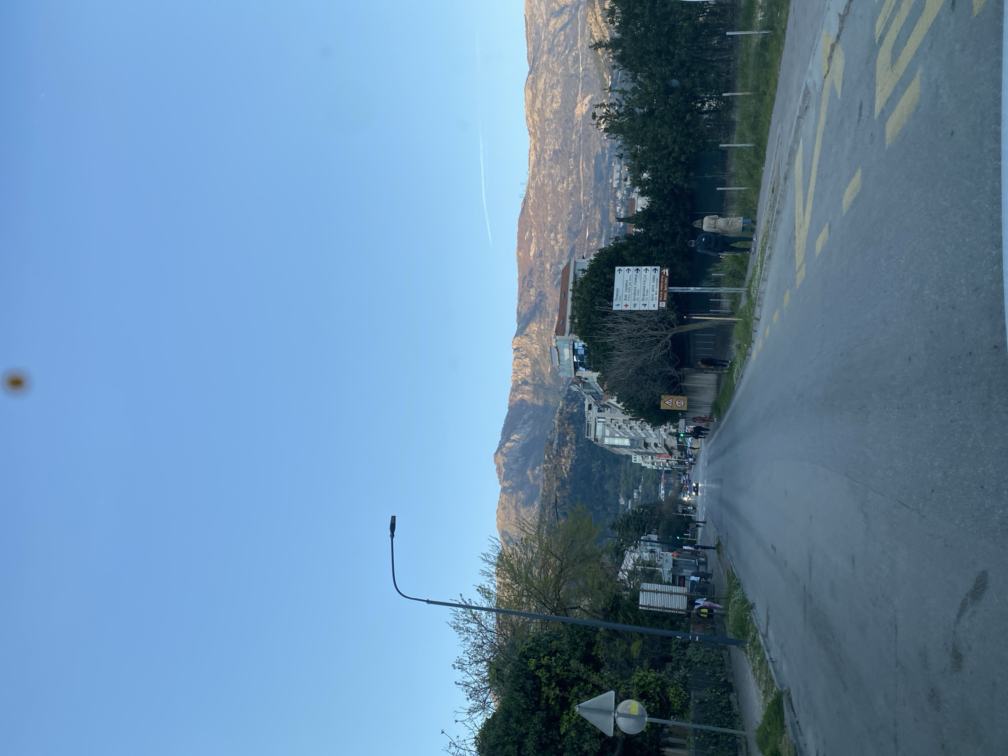 The image depicts a road in Budva, Montenegro, with a mountain range in the background. The sky is clear blue with a contrail visible. A street lamp is on the left side of the road. There are trees and vegetation along the roadside. Buildings are visible along the mountain slope. The road has yellow markings. People are walking on the pavement. There are traffic signs.